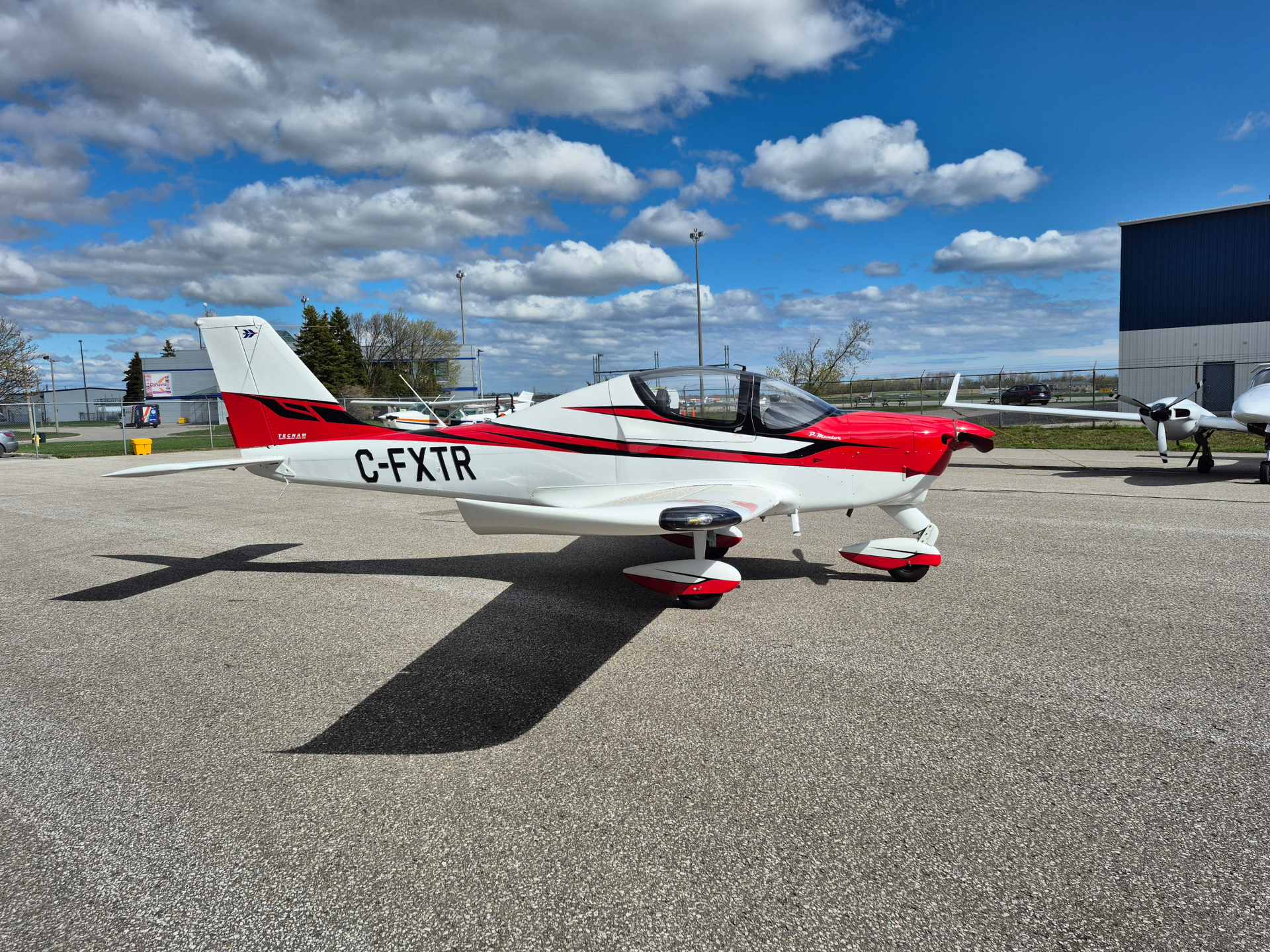 Small red-and-white single-engine aircraft parked on a tarmac with a blue sky and scattered clouds above, shadow visible on the ground.