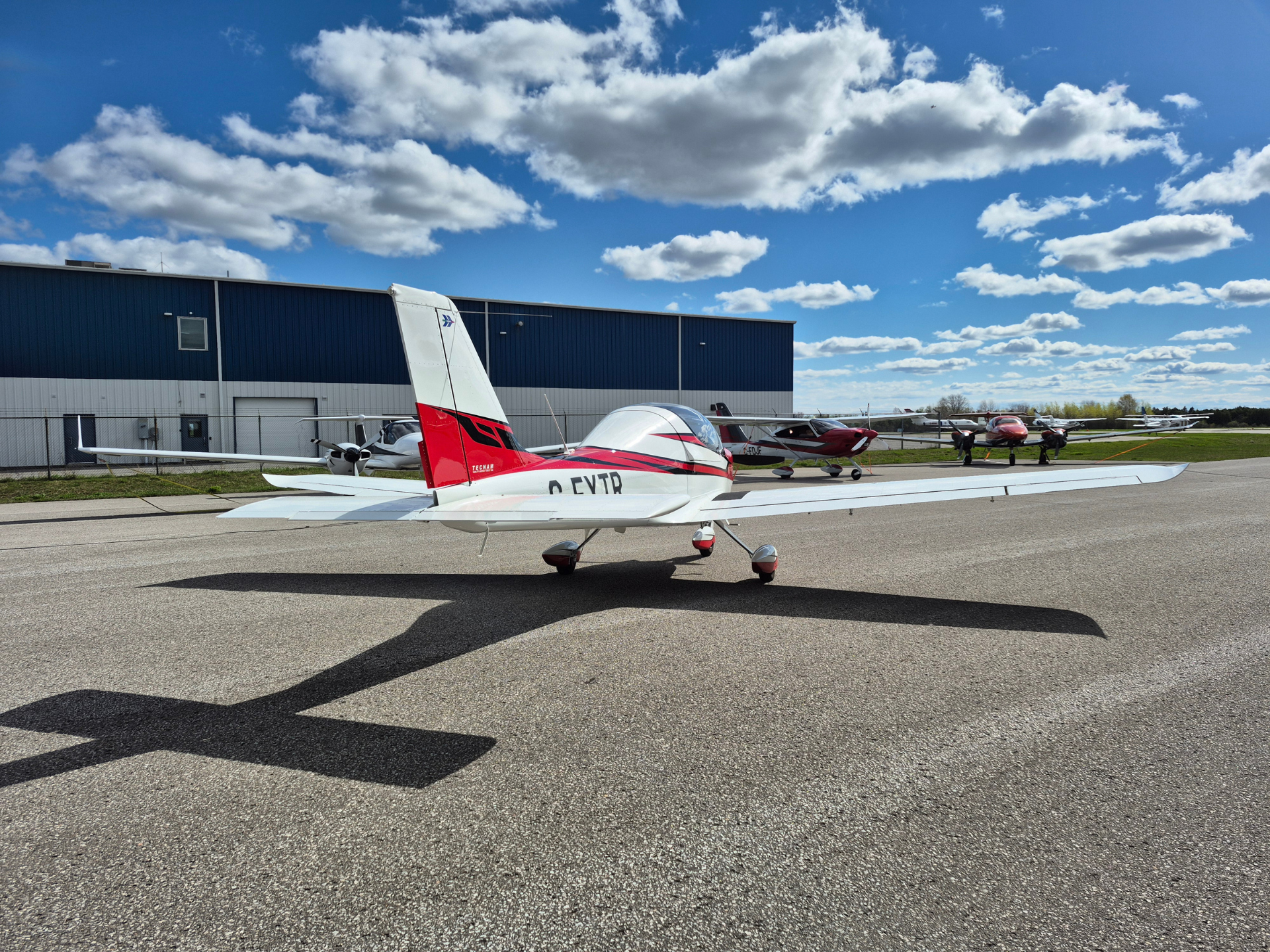 Small white and red single‑engine airplane on a tarmac, blue hangar behind, with other planes in the background and a bright sky.