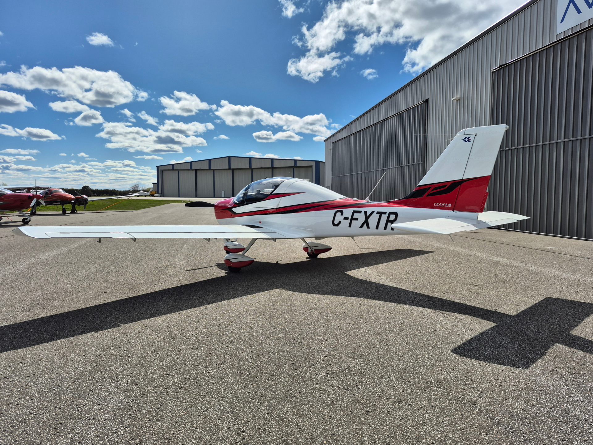 Small white and red single-engine airplane parked on a taxied ramp beside a metal hangar under a bright blue sky with scattered clouds.