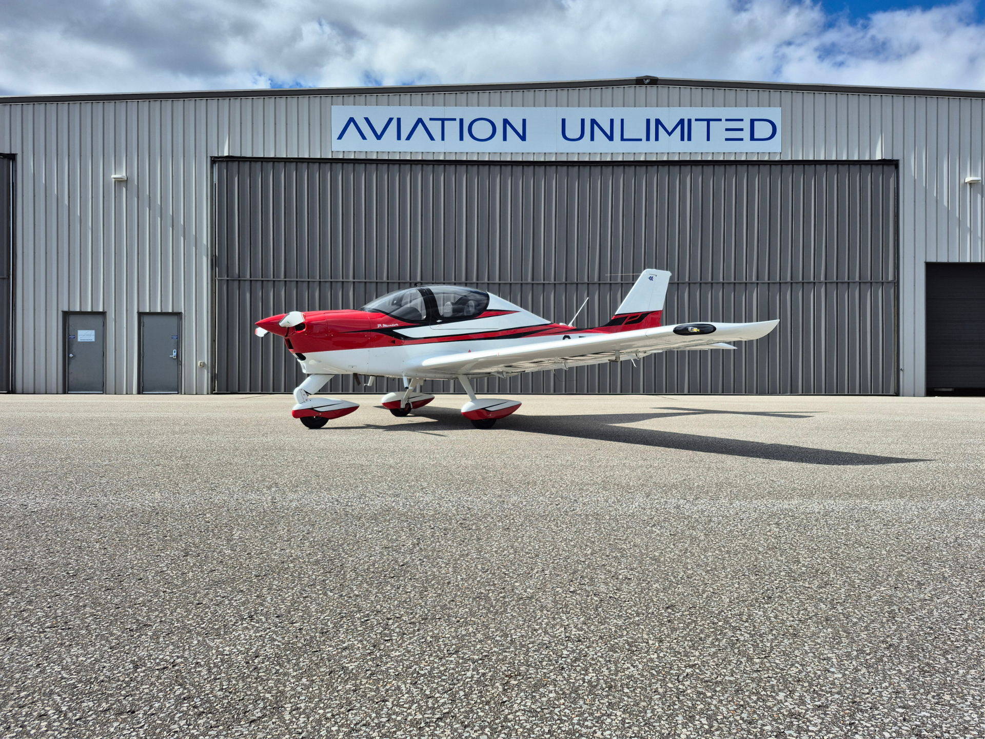 Small red-and-white single-engine airplane parked on a taxiway in front of a gray hangar with 'AVIATION UNLIMITED' sign.