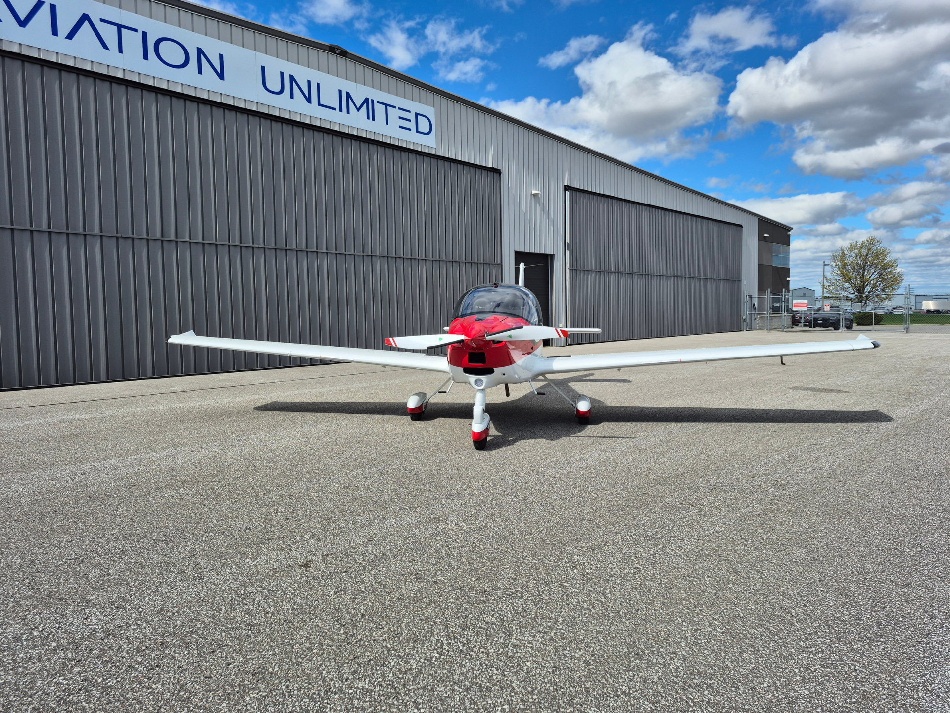 Small red-and-white single-engine airplane parked on a tarmac in front of a gray hangar under a blue sky with clouds.