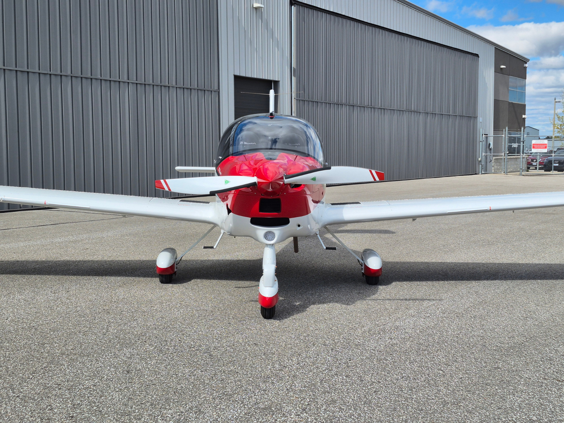 Small single-engine airplane facing the camera on a paved tarmac with a gray hangar in the background, red nose and white fuselage.