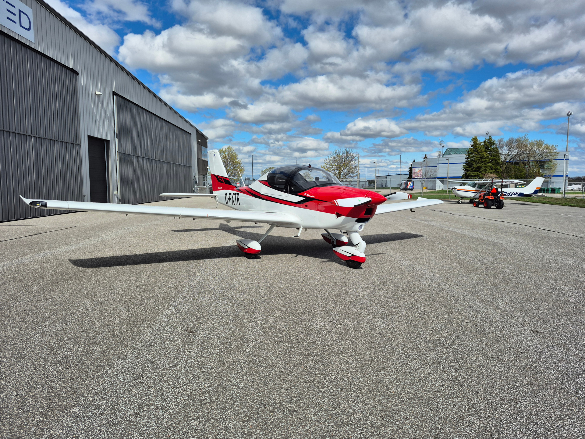 Small red-and-white airplane parked on a tarmac beside a gray hangar under a cloudy blue sky.