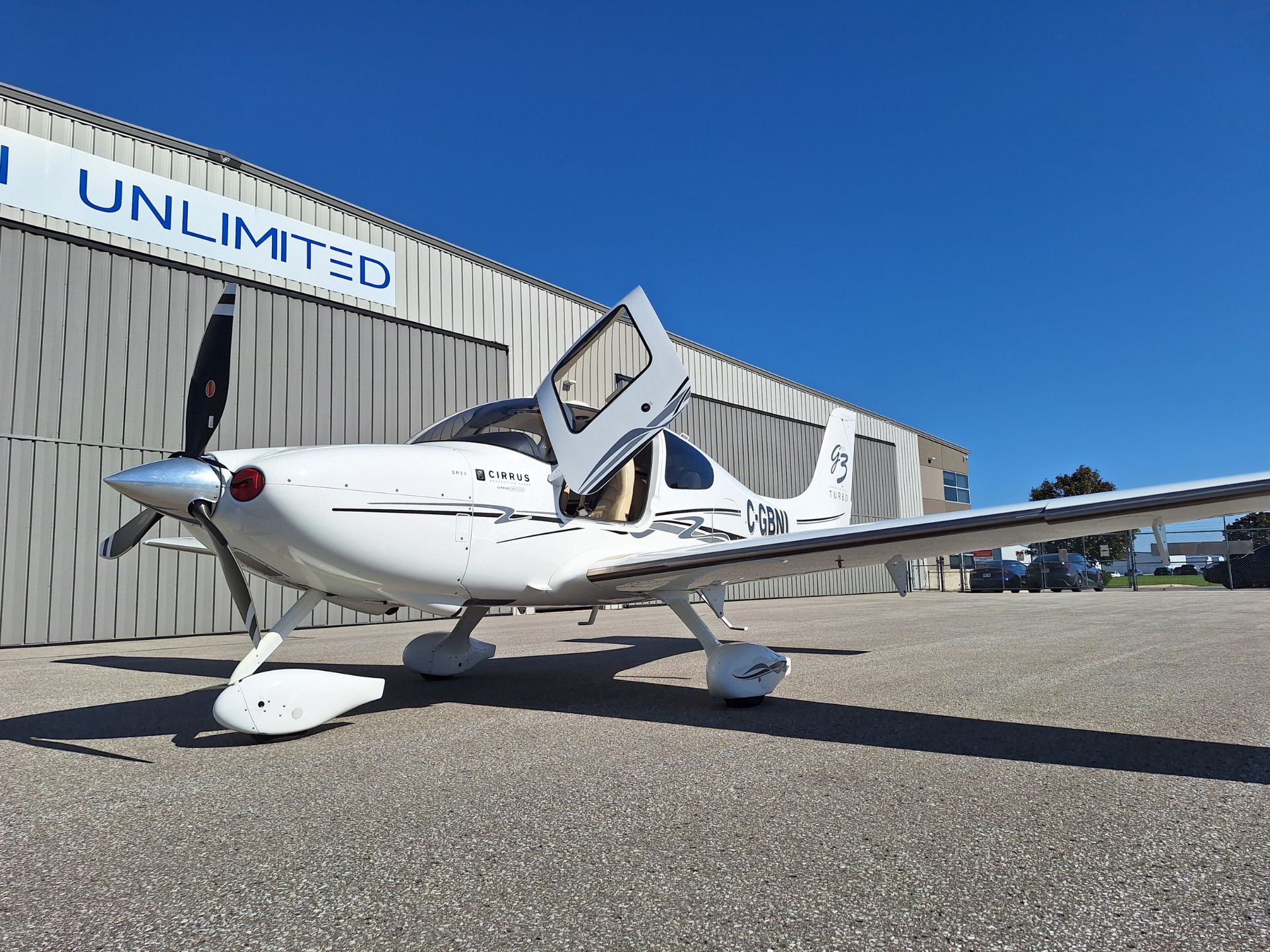 Small white single-engine airplane on tarmac in front of a gray hangar with a blue 'UNLIMITED' sign against a clear blue sky.