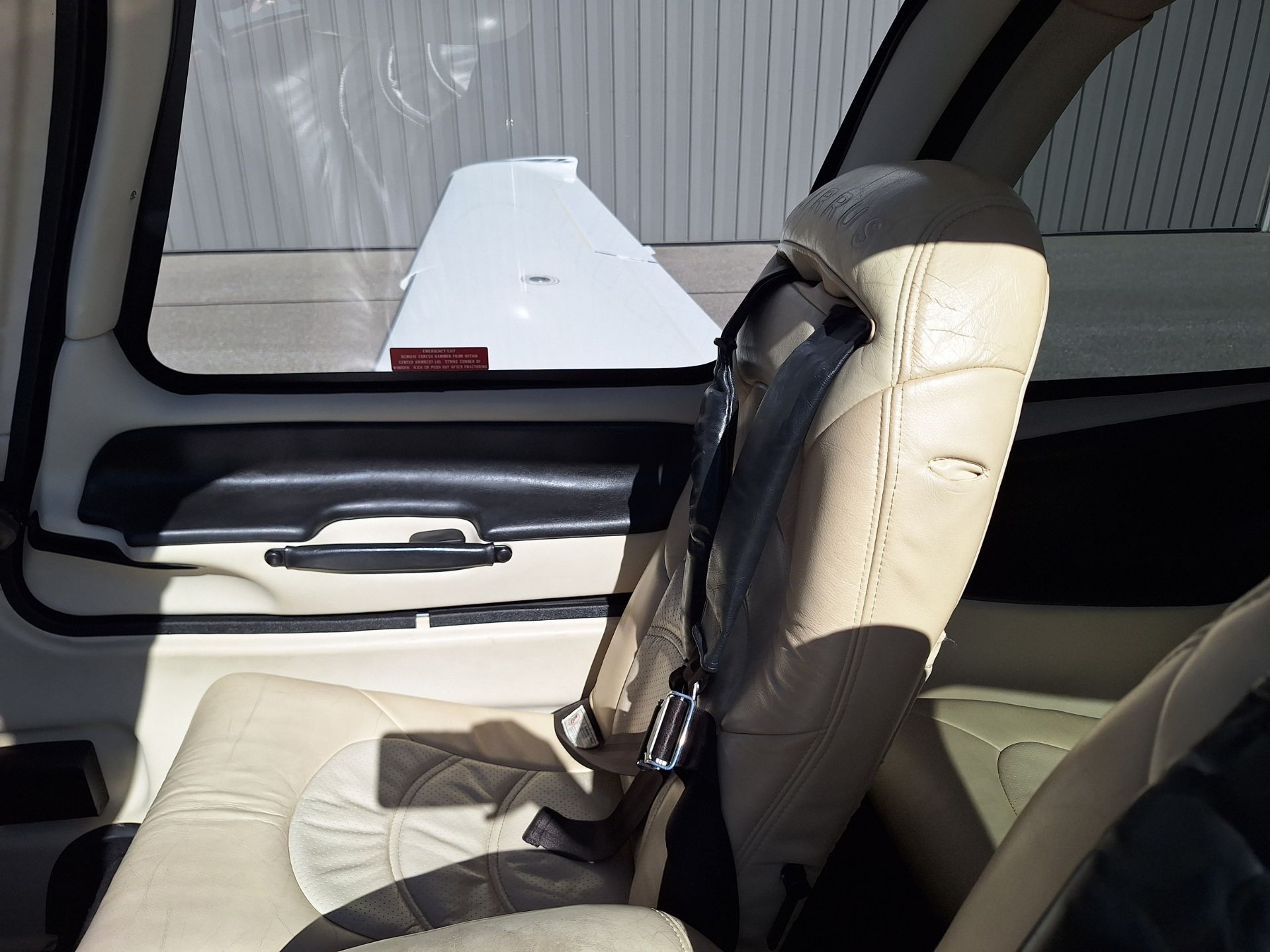 Beige aircraft cabin seat with a seatbelt; window shows the wing outside a hangar.
