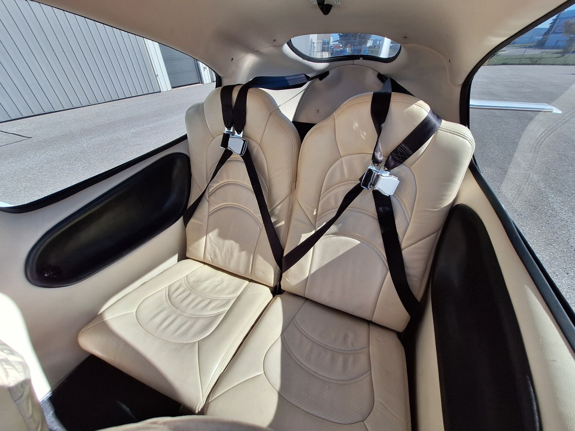 Two beige leather airplane seats with black shoulder restraints in a small cabin, window on right side showing tarmac outside