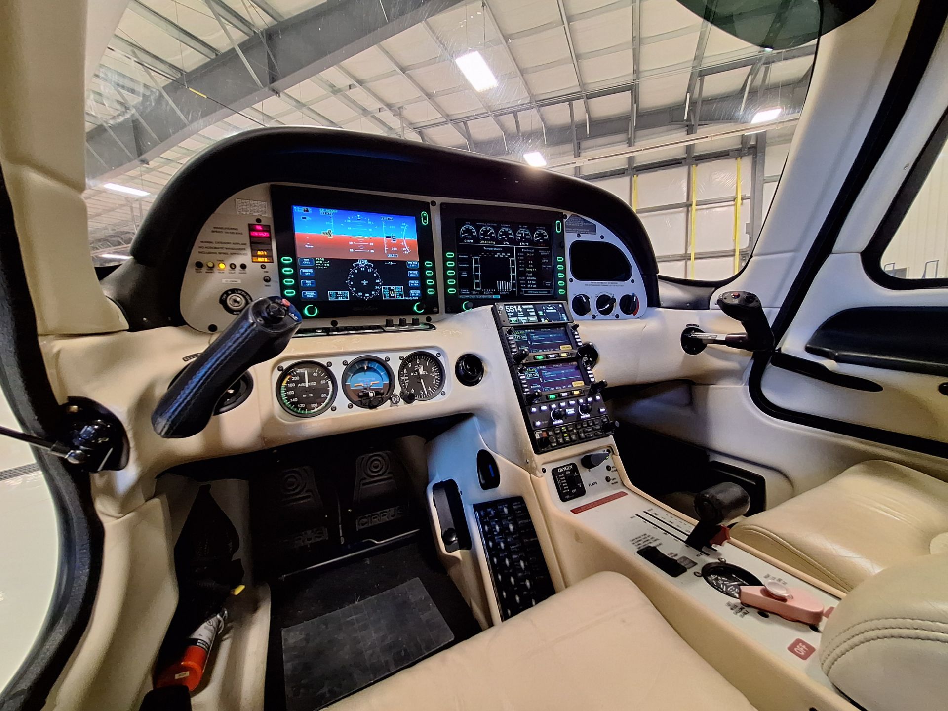 Pilot cockpit with dual large displays, analog gauges, and a center smartphone-like tablet mounted; hangar visible through windshield