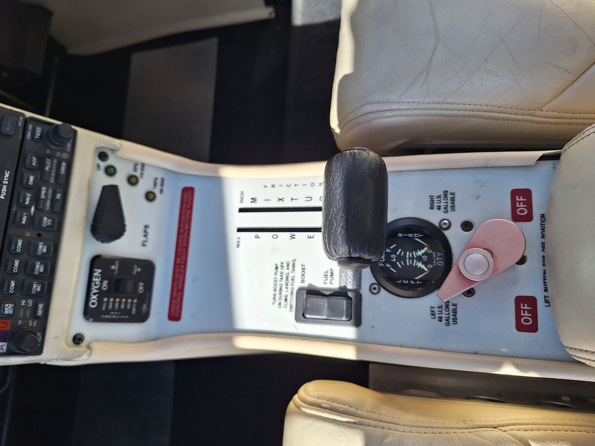 Center cockpit console with throttle levers and various switches, beige seat nearby, cockpit controls visible in sunlit cabin