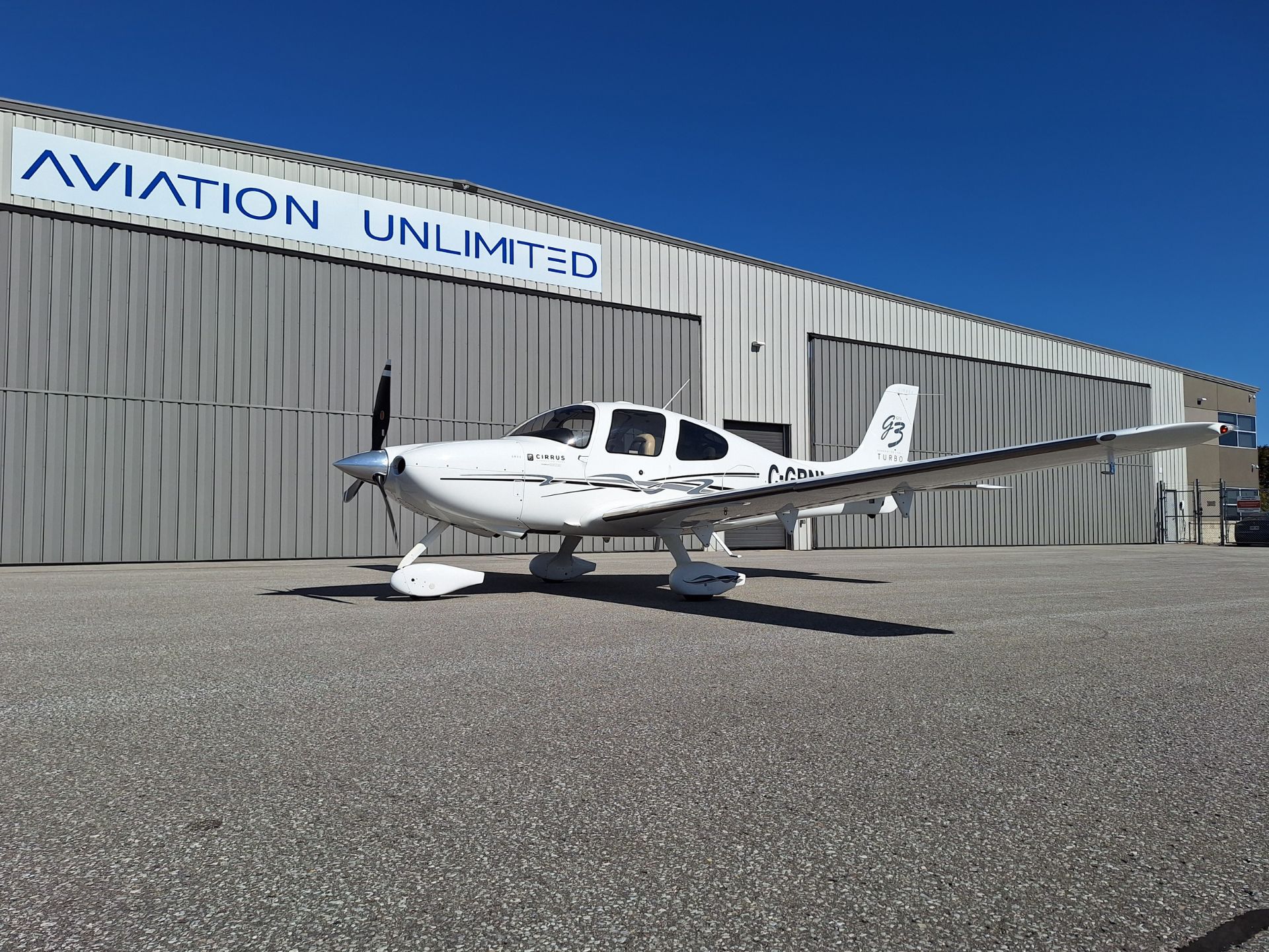 White Cirrus turbo aircraft parked on concrete tarmac in front of a gray metal hangar with an 'AVIATION UNLIMITED' sign against a clear blue sky.