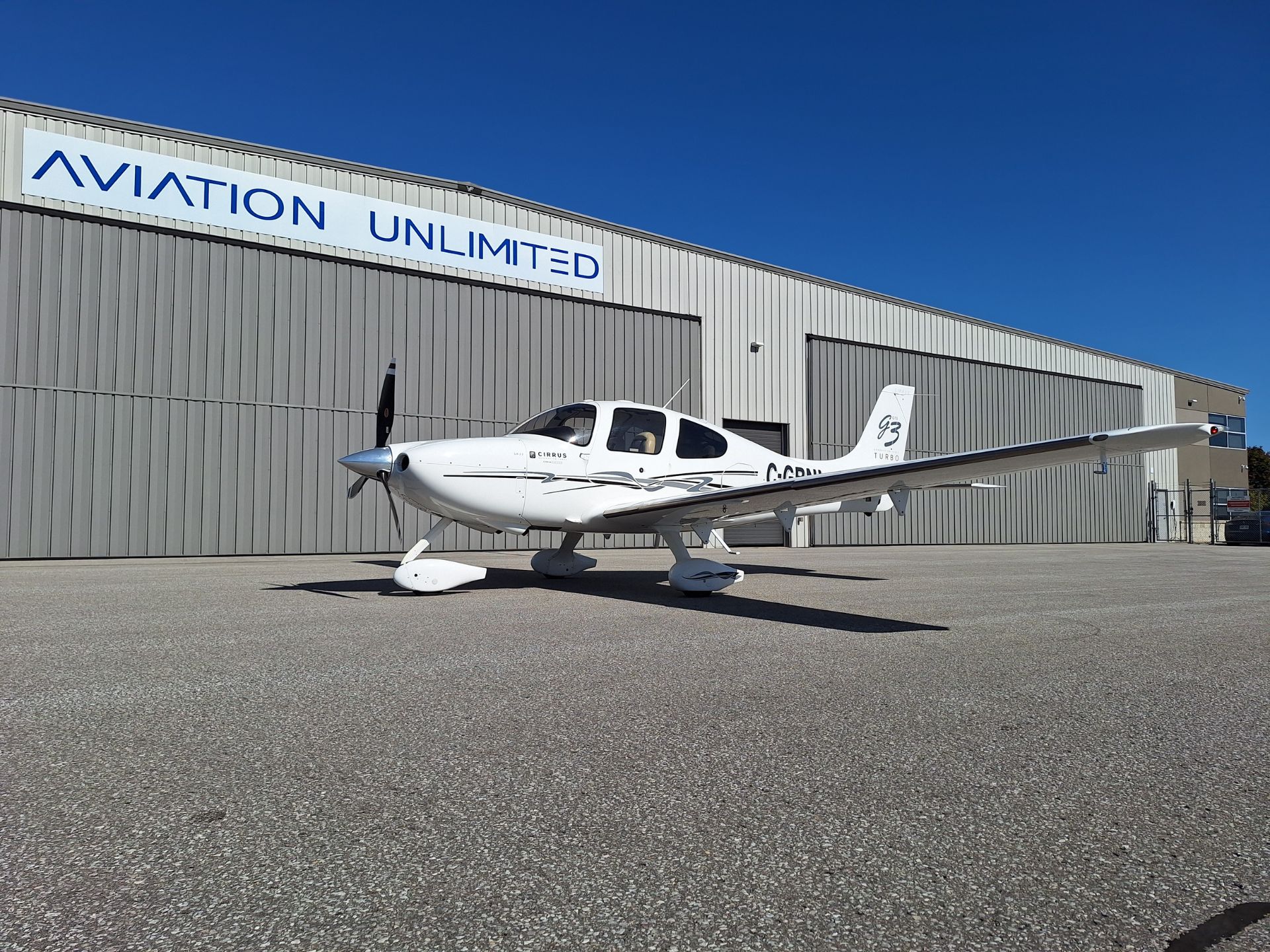 White Cirrus turbo airplane on the tarmac in front of a gray metal hangar with a blue sky and the sign 'AVIATION UNLIMITED' above.