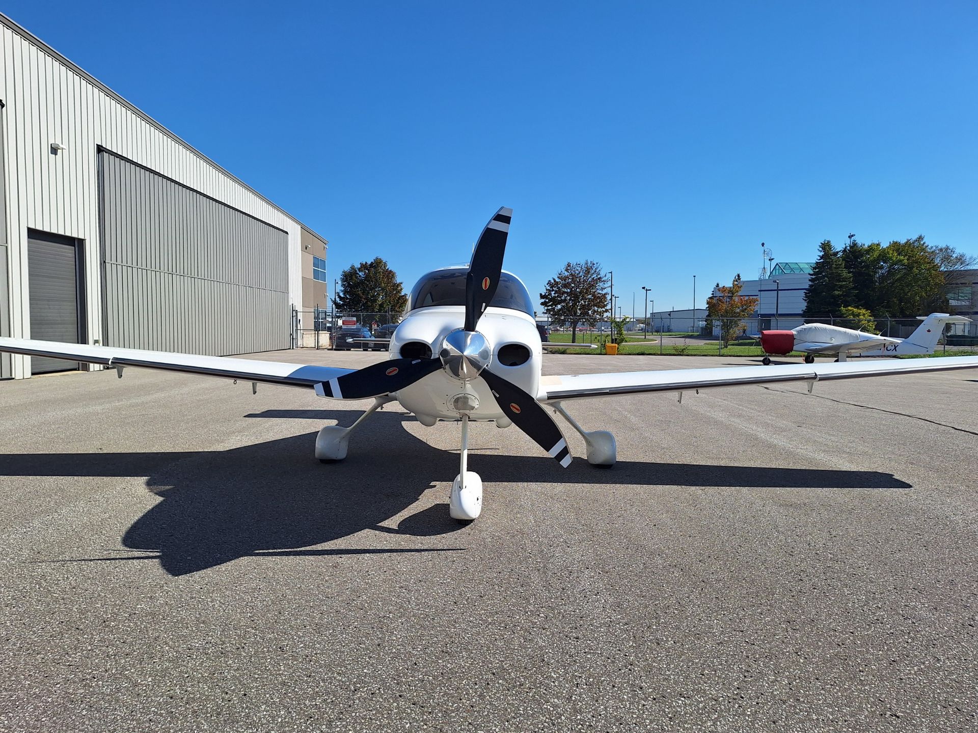 Small white single-engine airplane parked on tarmac with blue sky and hangar to the left; another plane in background beyond fence.