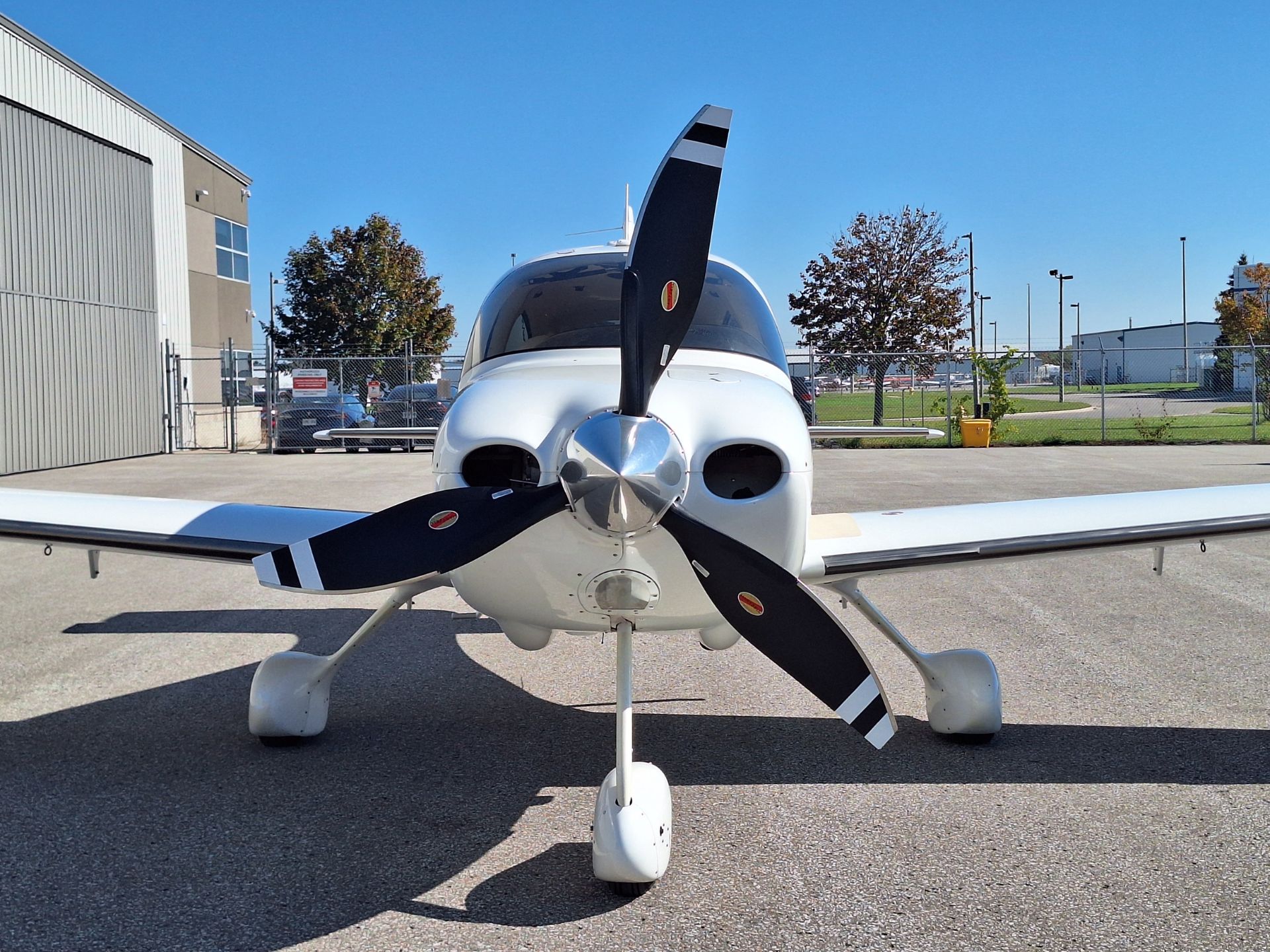 Front view of a white single-engine propeller airplane parked on a tarmac, with a hangar and chain-link fence in the background under a clear blue sky.
