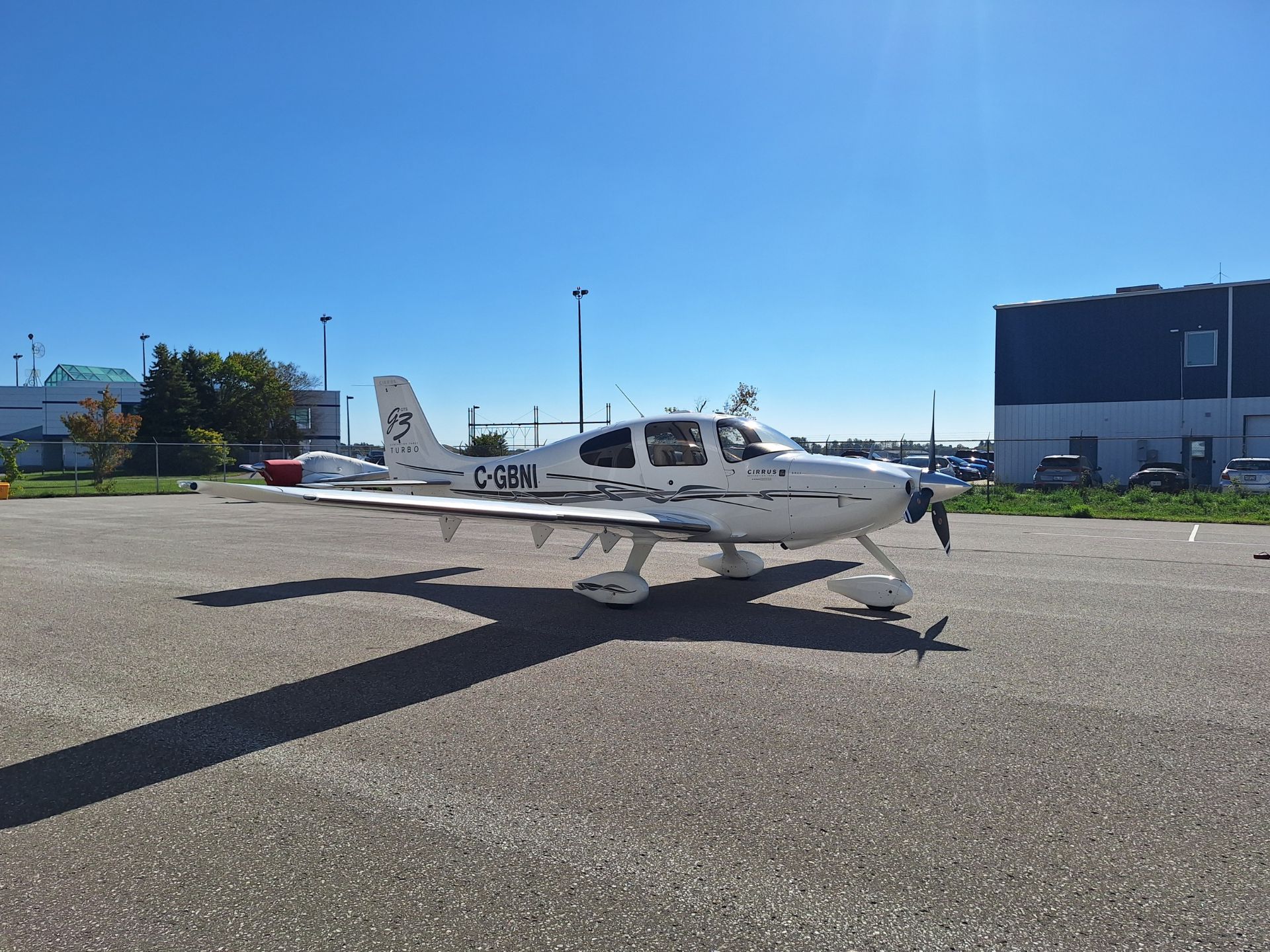 White Cirrus single-engine airplane parked on a sunlit tarmac, tail number C-GBNI, with blue sky overhead.