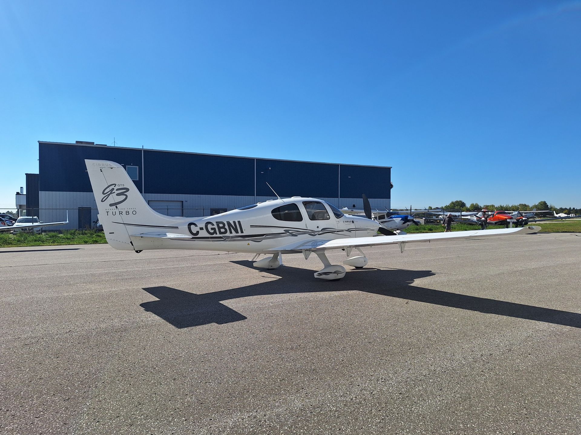 White Cirrus GTS Turbo small aircraft on tarmac with blue hangar in background, clear sky above