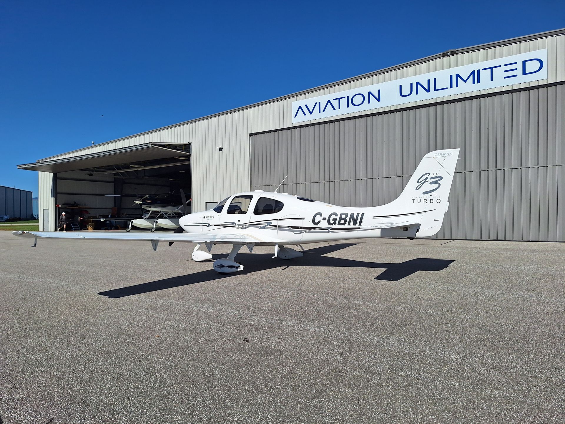 Small white Cessna-style airplane parked outside a hangar labeled Aviation Unlimited, with a blue sky above.