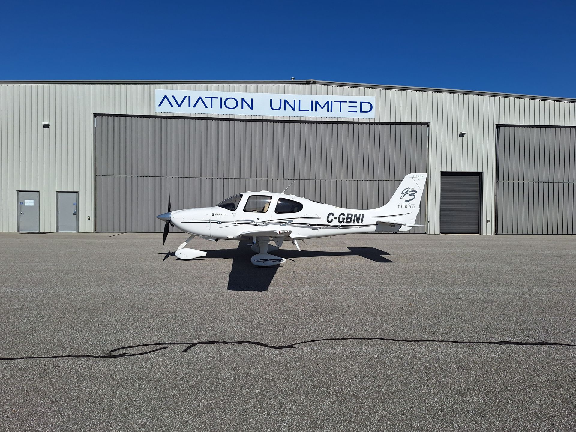 White Cirrus single-engine aircraft parked on the tarmac in front of a gray hangar with a blue 'AVIATION UNLIMITED' sign and a clear blue sky behind it.