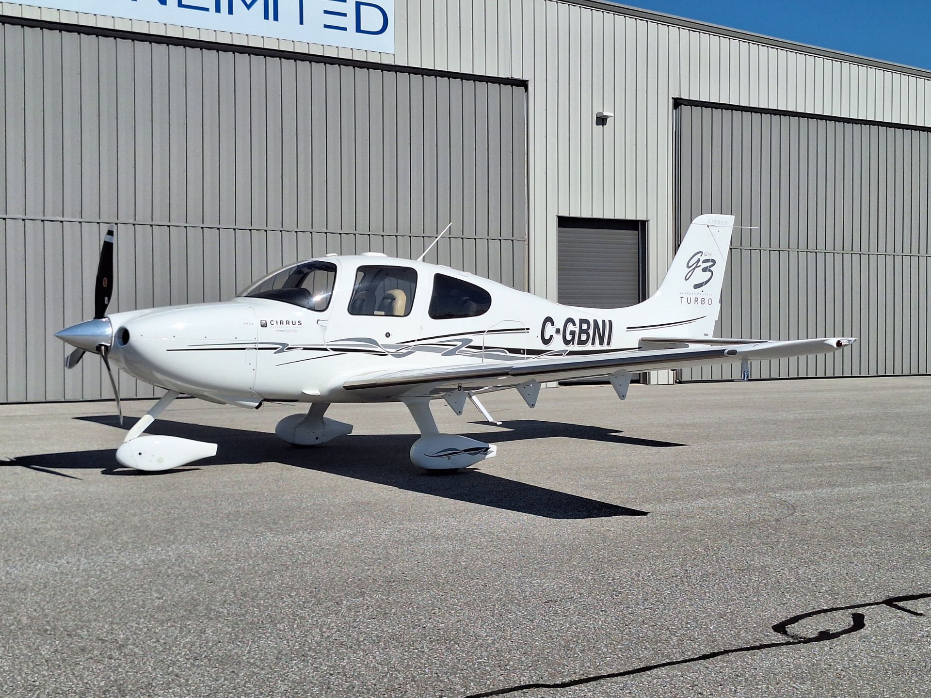 White Cirrus single-engine airplane parked on a tarmac in front of a gray metal hangar, registration C-GBNI, blue sky above.