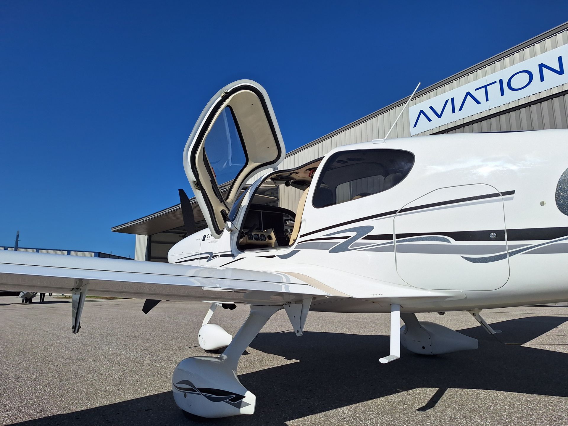 Small white private airplane on the tarmac with the cabin door open, at an aviation hangar under a clear blue sky.