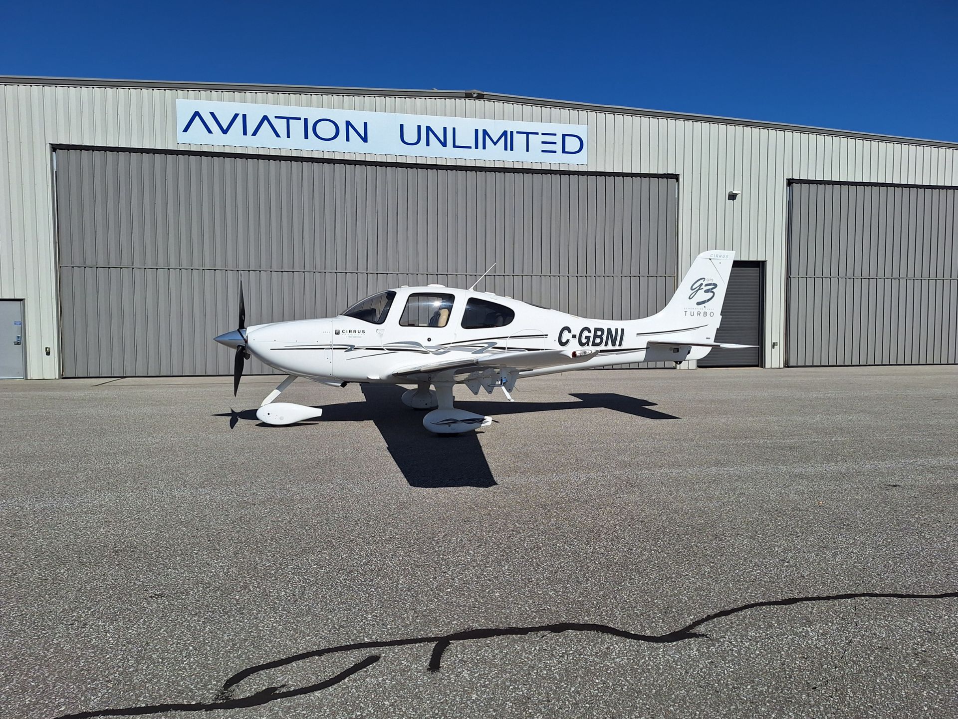 Small white Cirrus airplane parked on a tarmac in front of a gray hangar with a blue 'Aviation Unlimited' sign above.