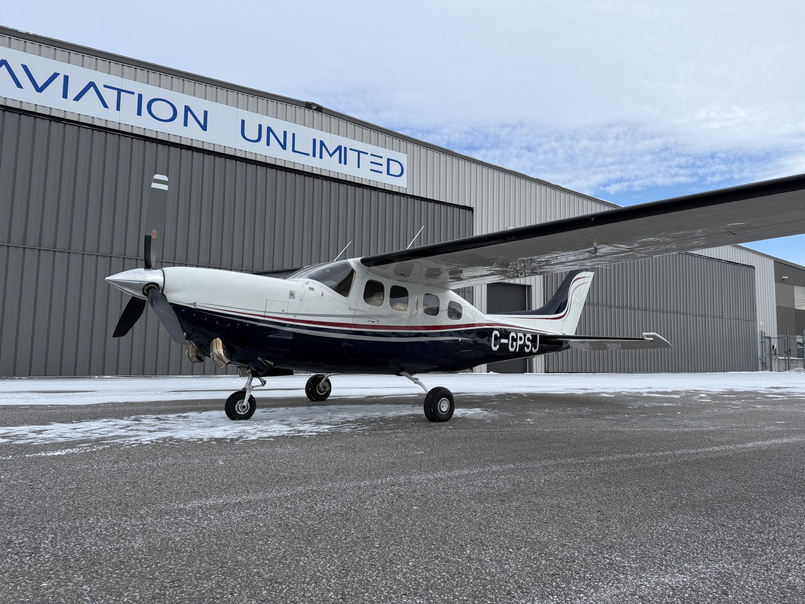 High wing single engine airplane on a snow dusted tarmac in front of a gray hangar with a large AVIATION UNLIMITED sign registration C GPSJ visible on fuselage