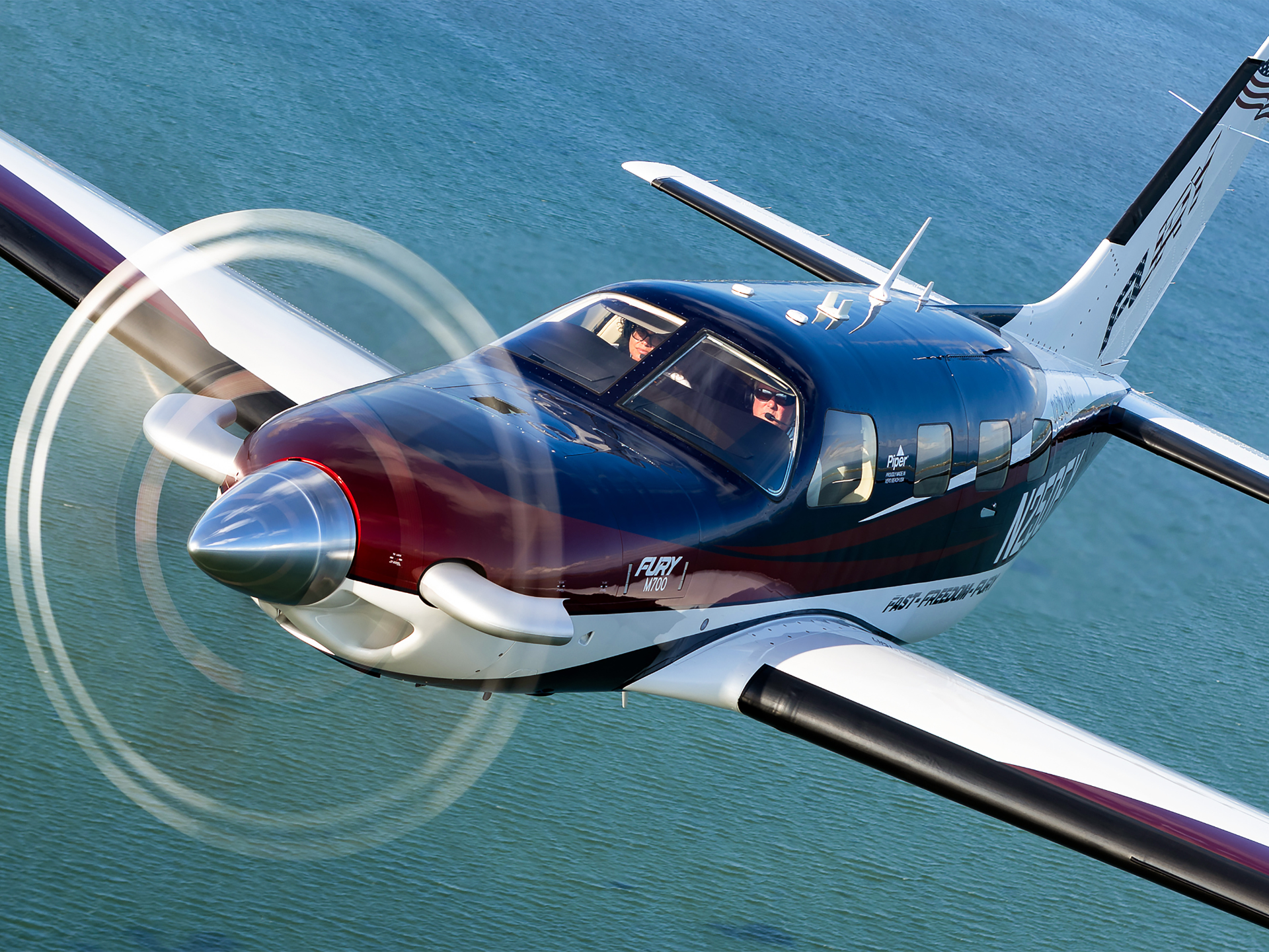 Blue and red twin-seat turboprop airplane in flight over calm blue water, cockpit windows showing two pilots.