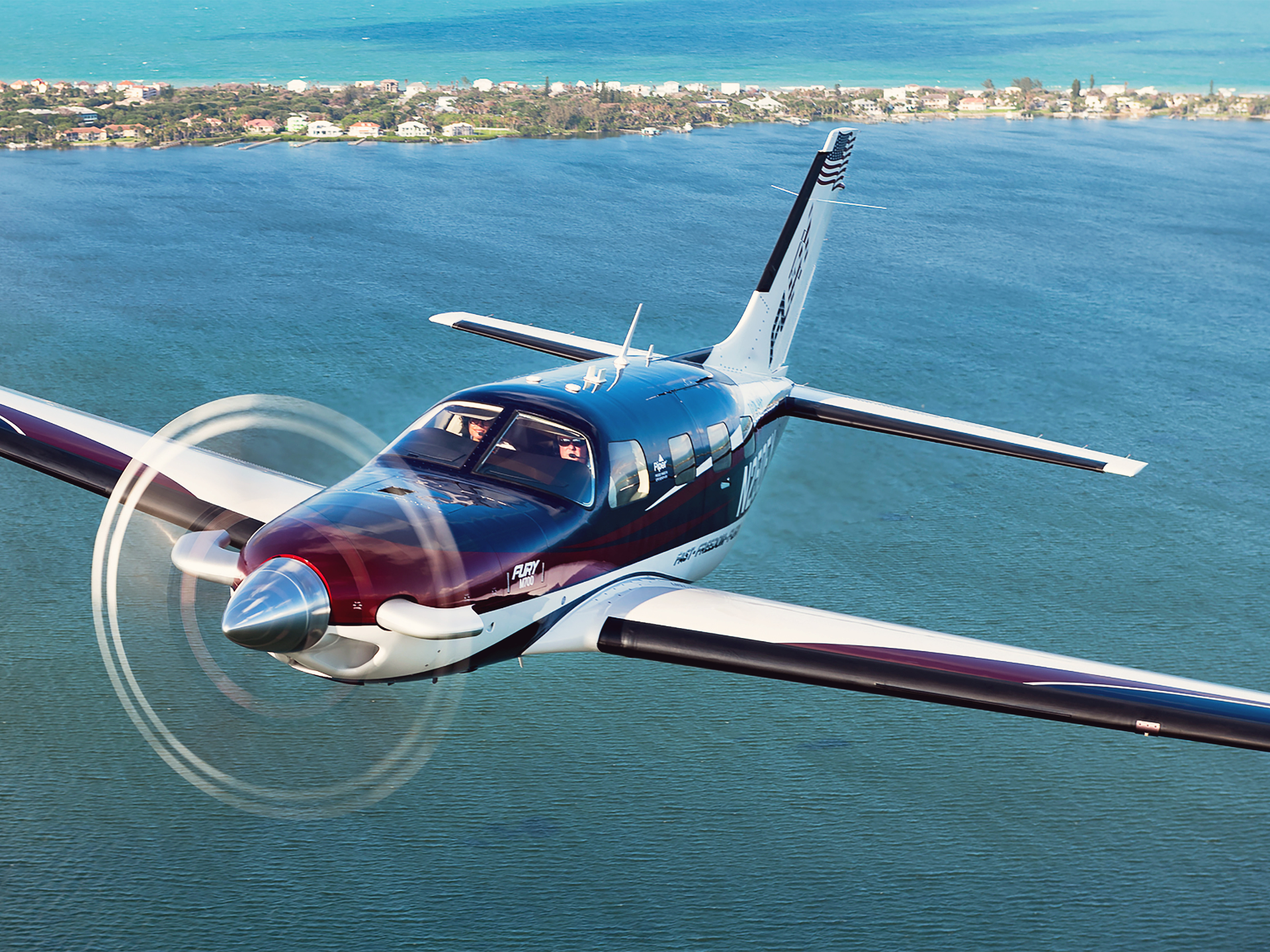 Single-engine propeller airplane with two pilots in the cockpit, flying over blue-green sea with a coastal town visible in the distance.