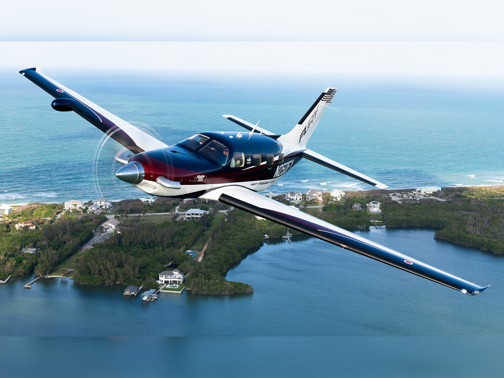 Single-engine airplane with long wings flying over a coastal town and blue ocean below.
