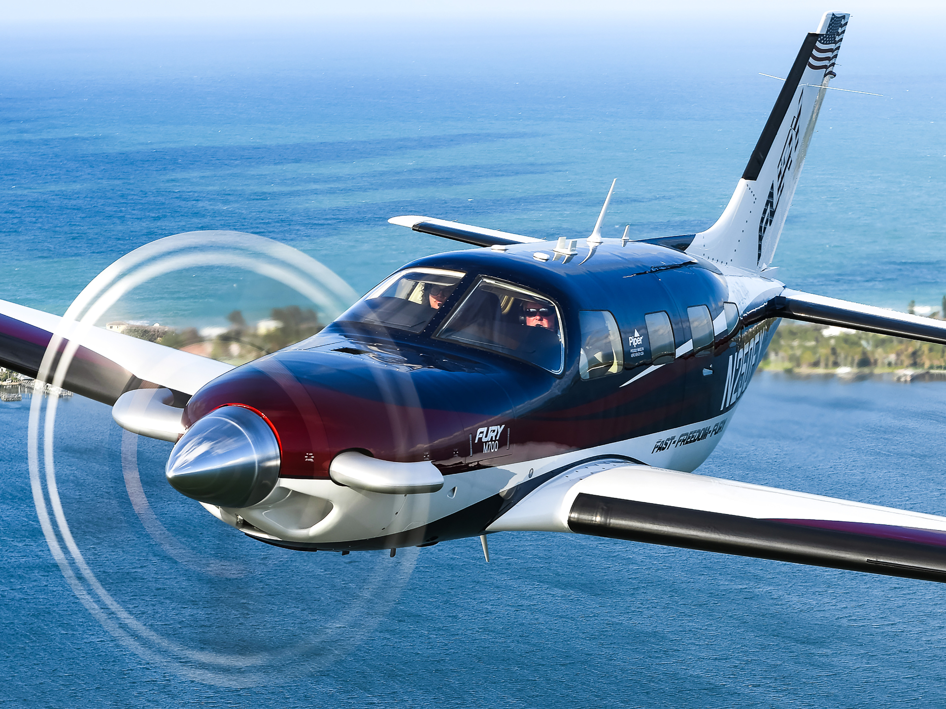 Blue and white light aircraft in flight over the ocean, propeller spinning with a coastline below.