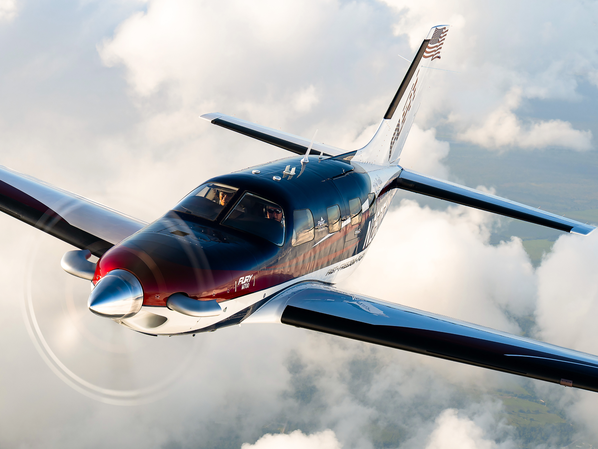 Piper M700 private airplane in dark blue and red livery flying above clouds, pilot visible in cockpit window with propeller spinning.
