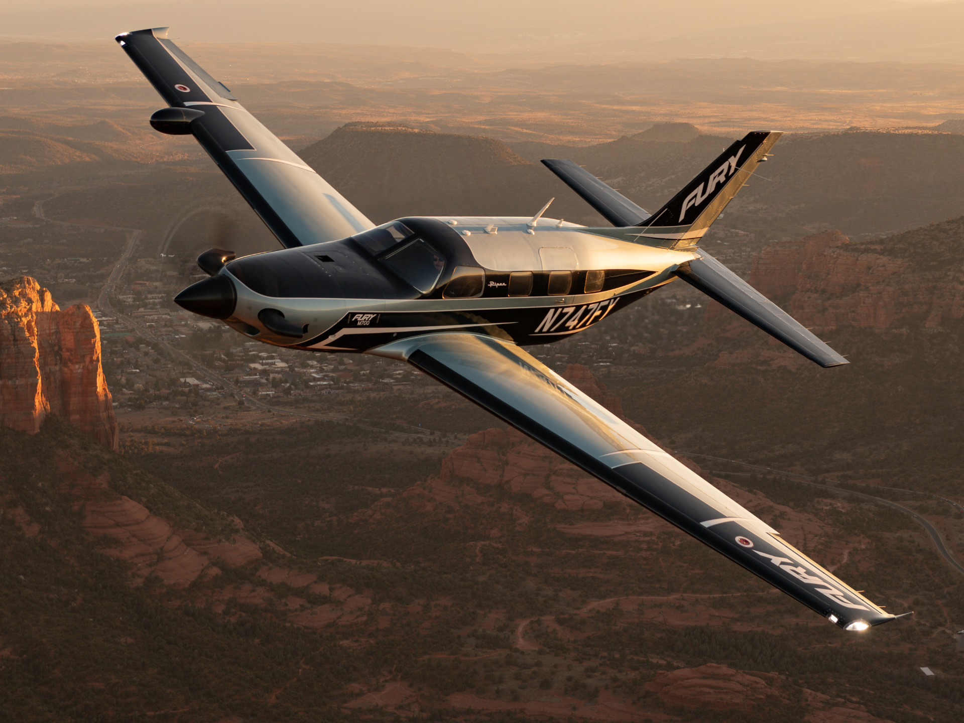 Piper aircraft flying over a desert canyon at sunset, with warm golden light on red rock formations below.