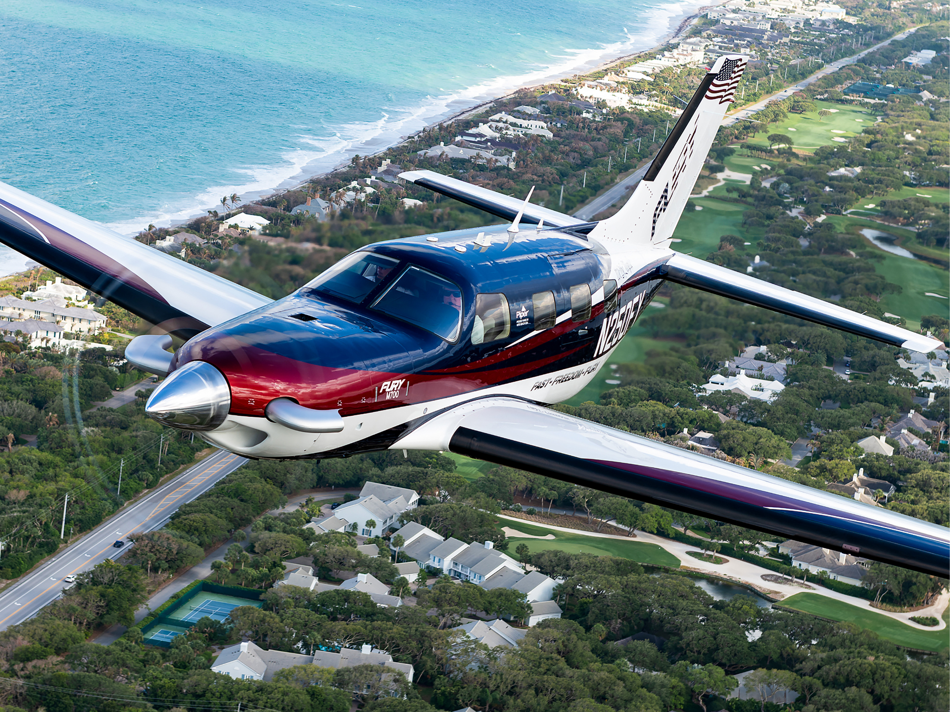 Small propeller airplane flying over a coastal neighborhood with the ocean to the left and a golf course to the right.