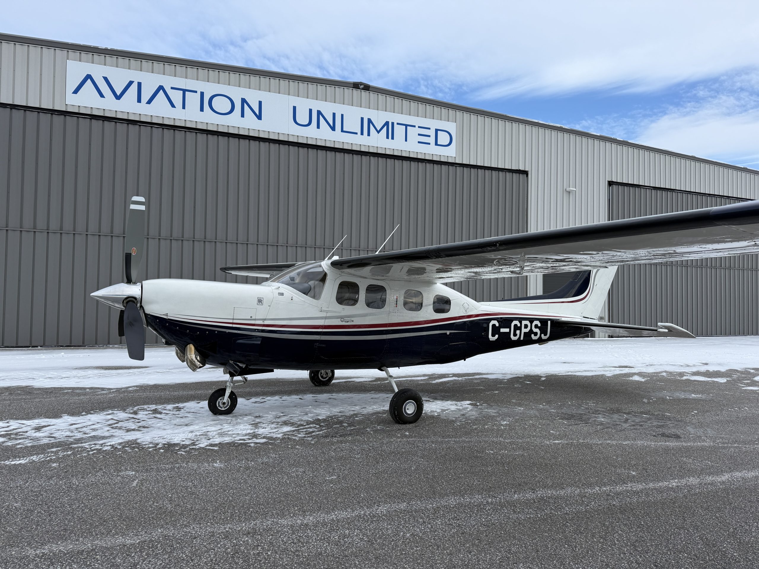 Small white and navy propeller airplane parked on a snow dusted tarmac in front of a grey hangar with an AVIATION UNLIMITED sign outside