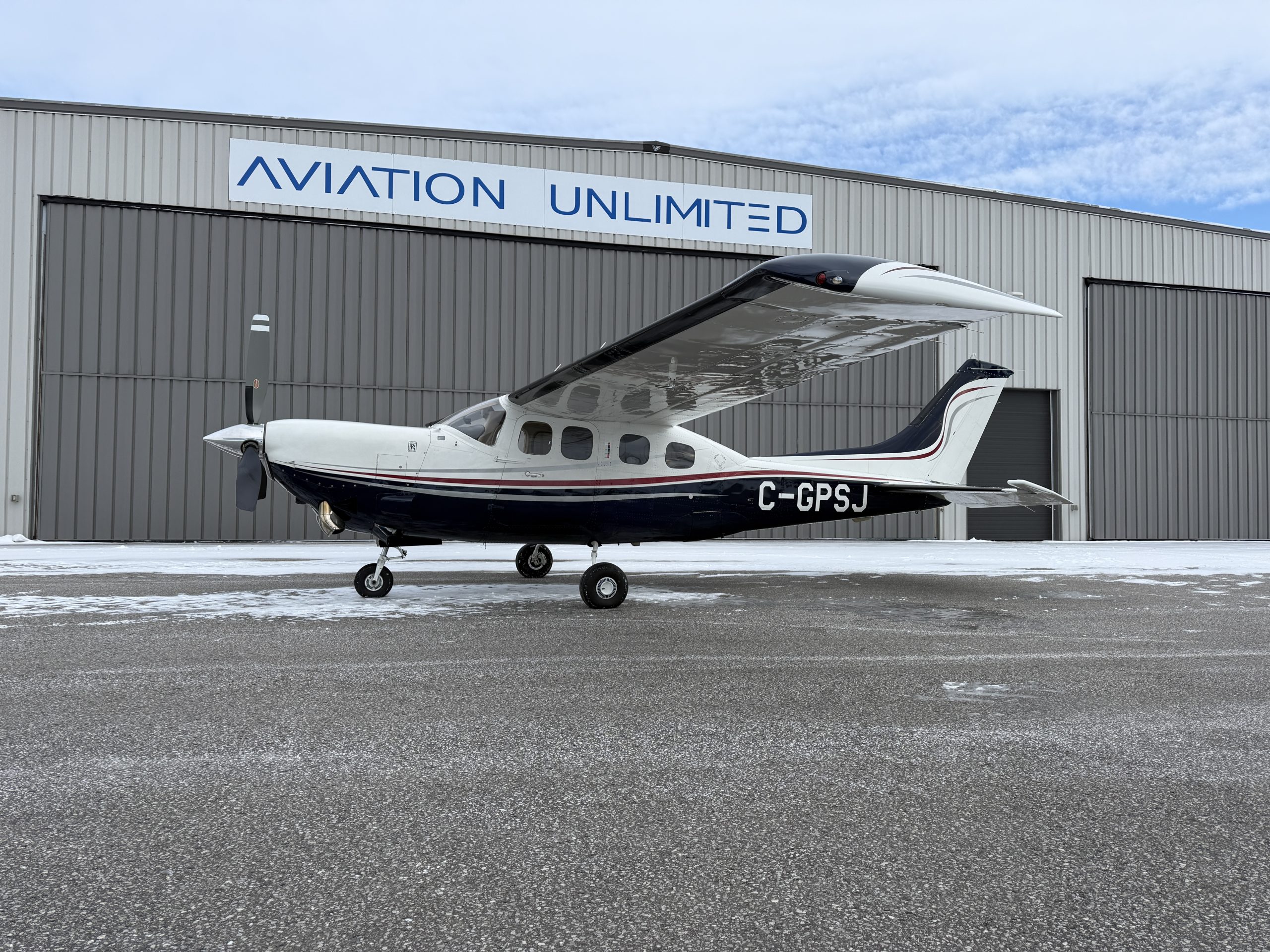Small propeller airplane parked in front of a gray hangar with an Aviation Unlimited sign on the building edge