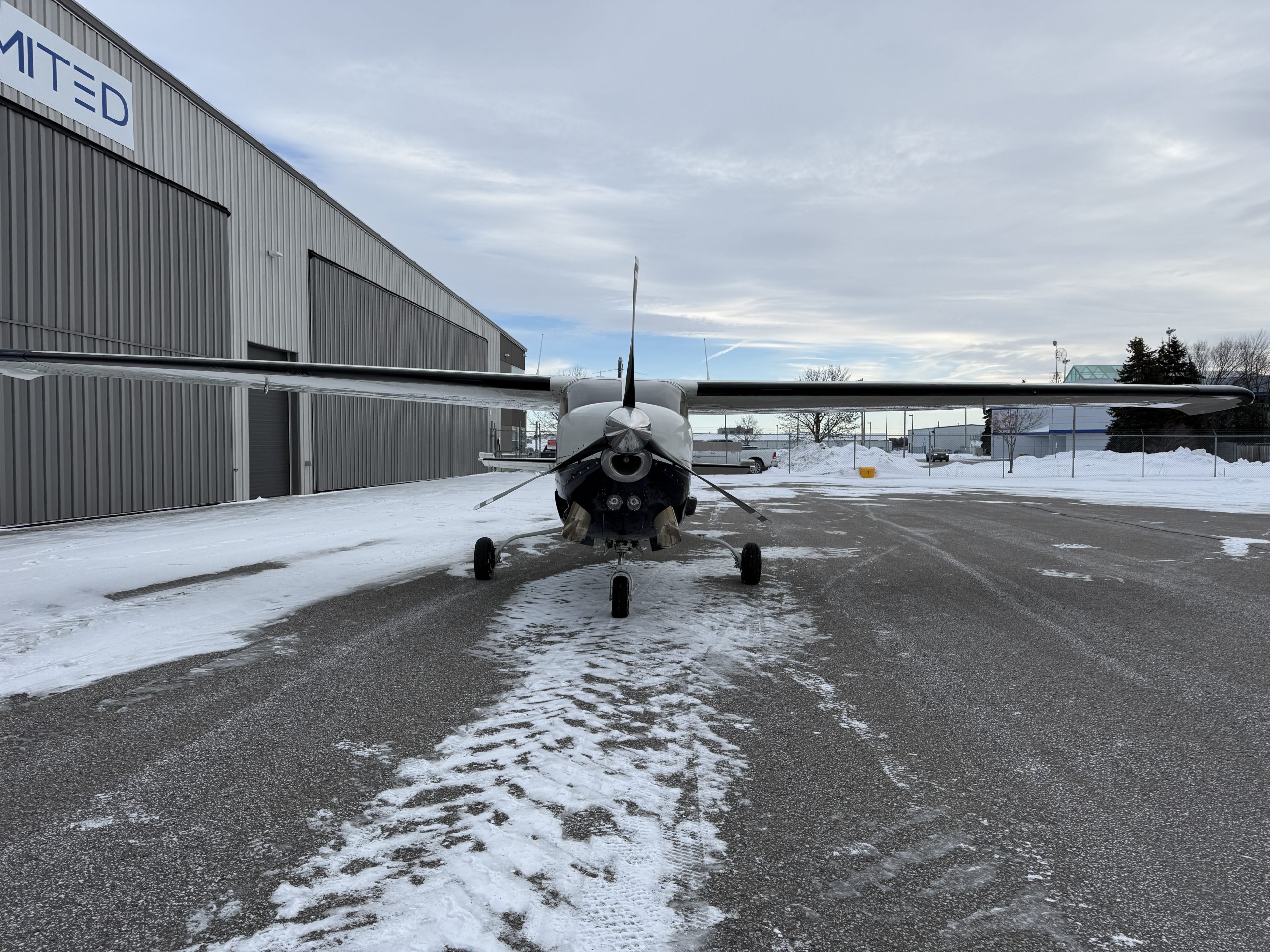 Front view of a small propeller airplane parked on a snow covered tarmac beside a gray industrial hangar