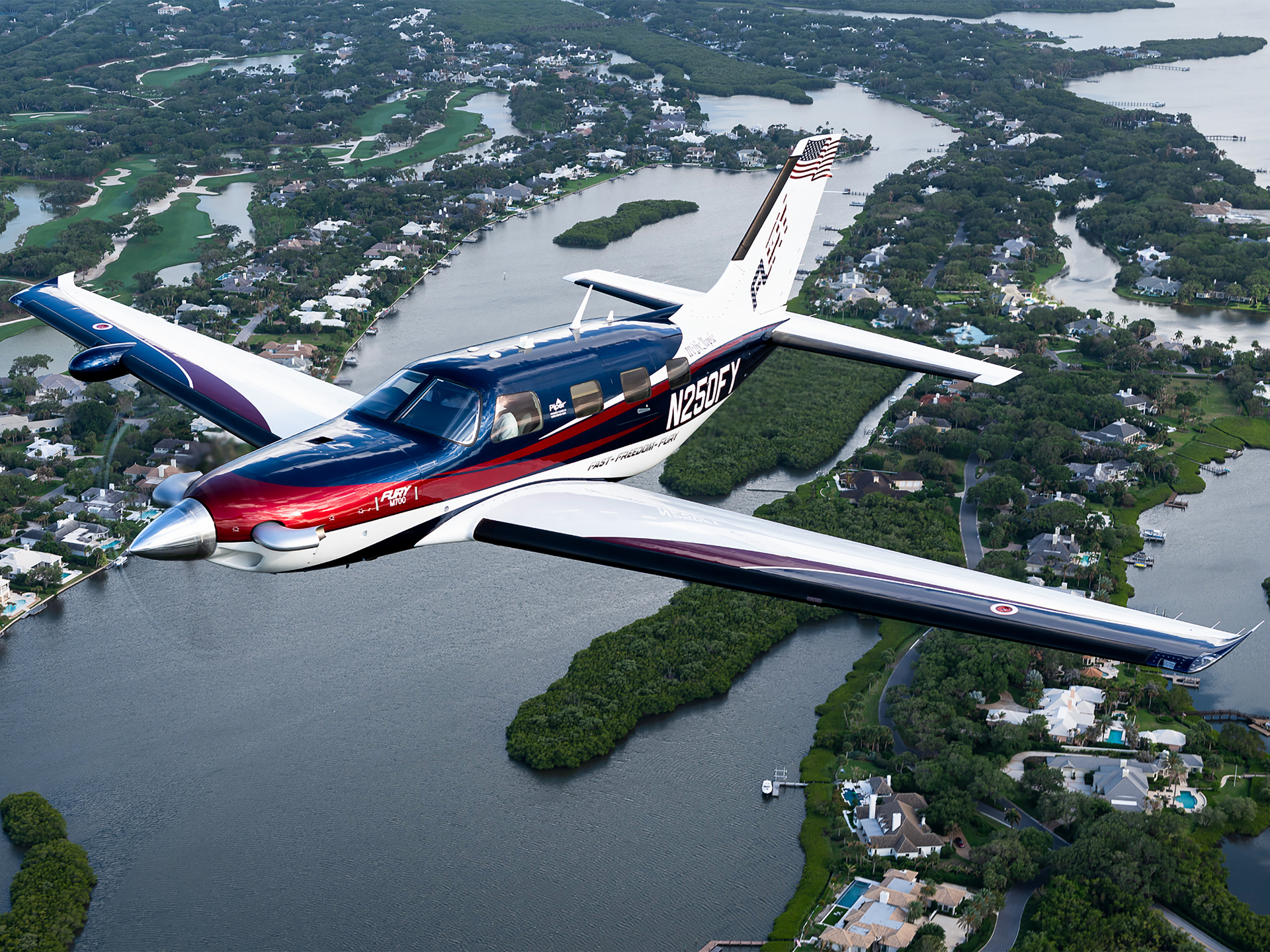Private single-engine airplane in blue, white, and red livery flying over a canal-front residential area.