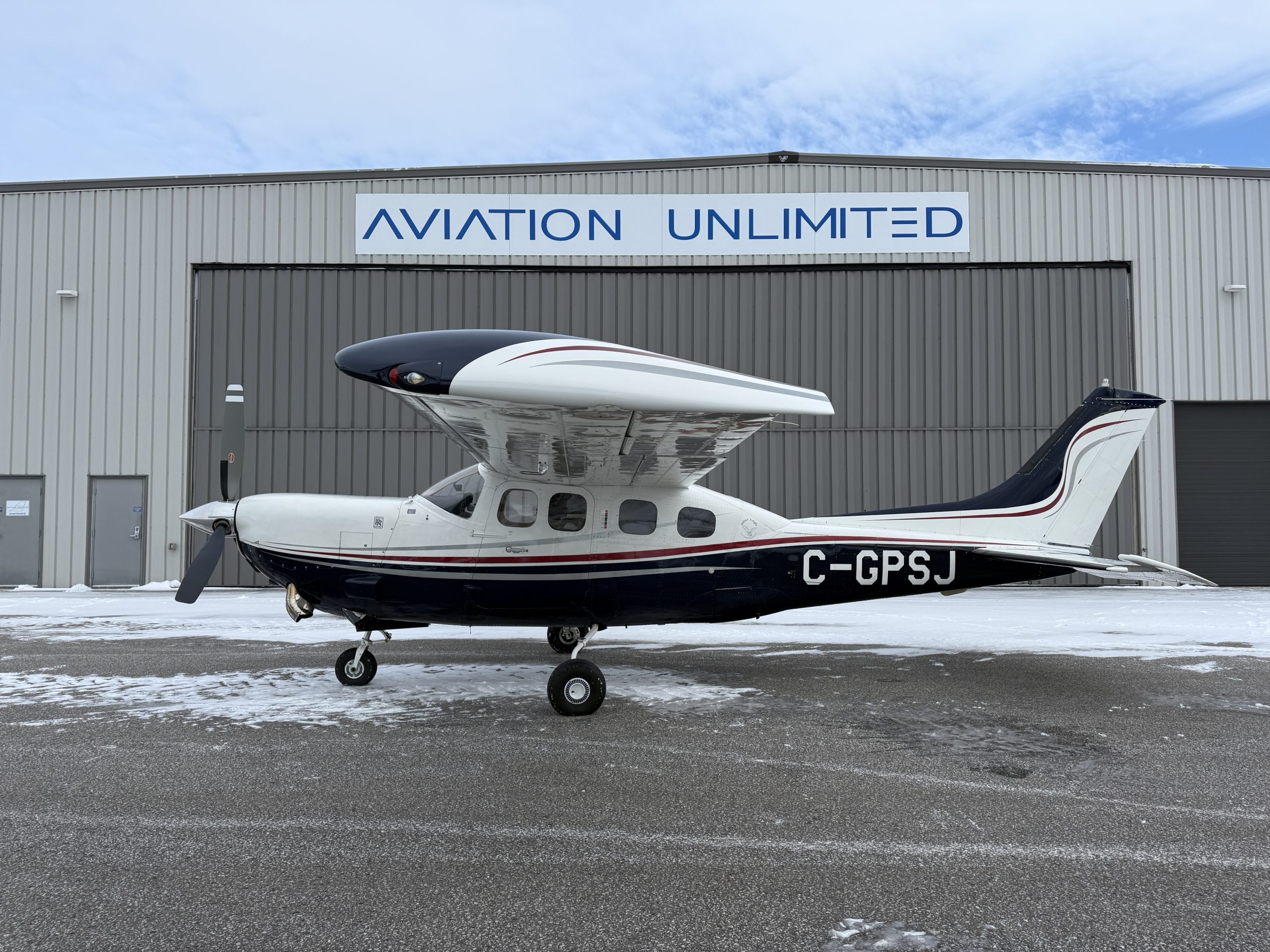 Small single engine airplane parked in front of a gray hangar with a blue AVIATION UNLIMITED sign above Swift tailed propeller plane on snow