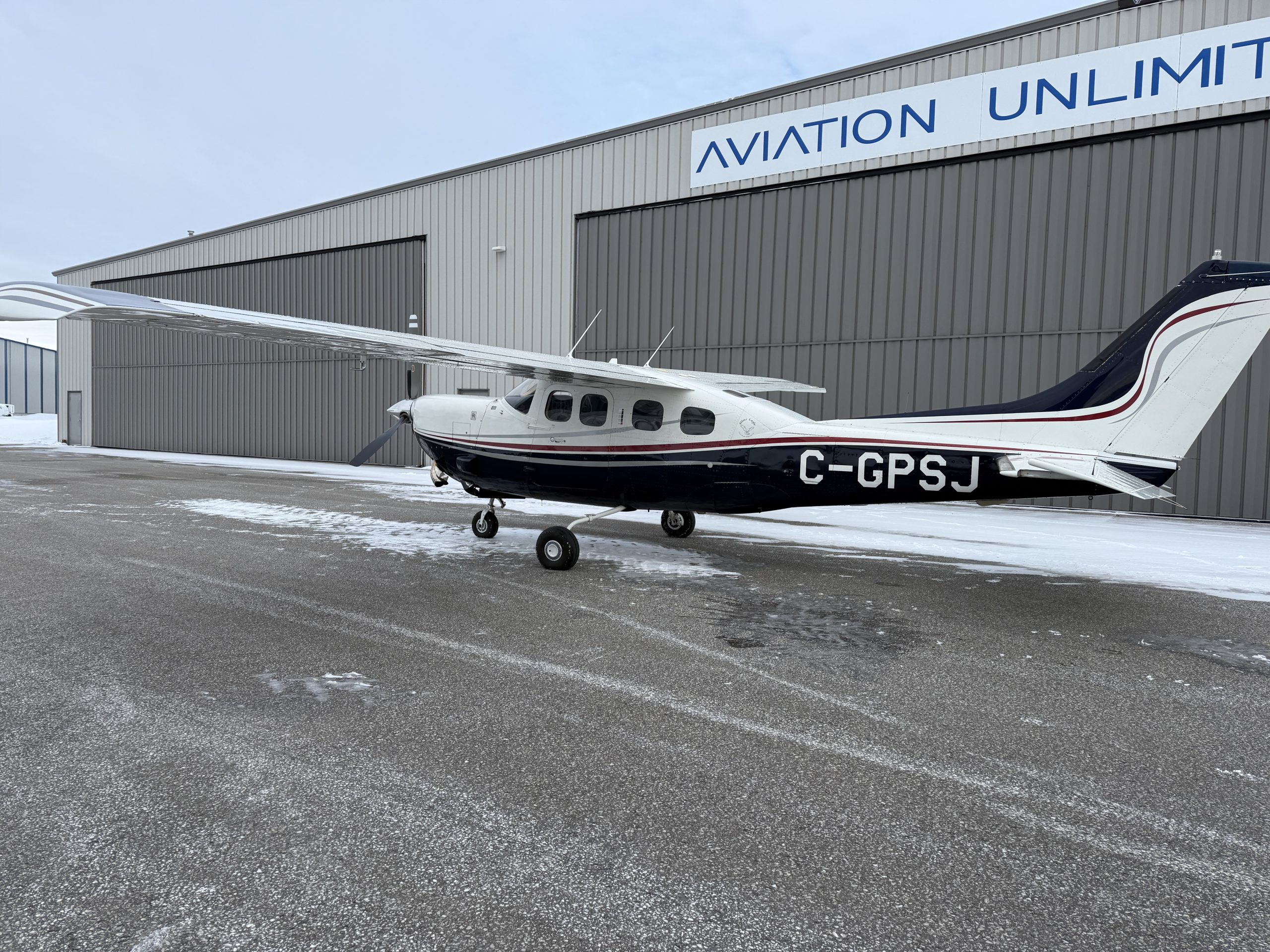 Small white and blue single engine plane parked on a snow dusted tarmac in front of a gray hangar with an Aviation Unlimited sign tail number C GPSJ visible