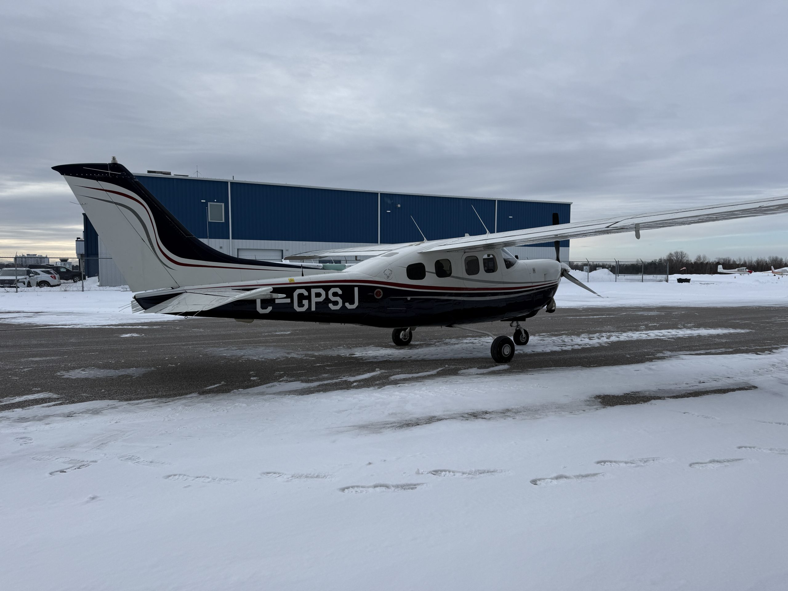 Small white and black propeller airplane on snow beside a blue hangar under an overcast sky at a winter airfield