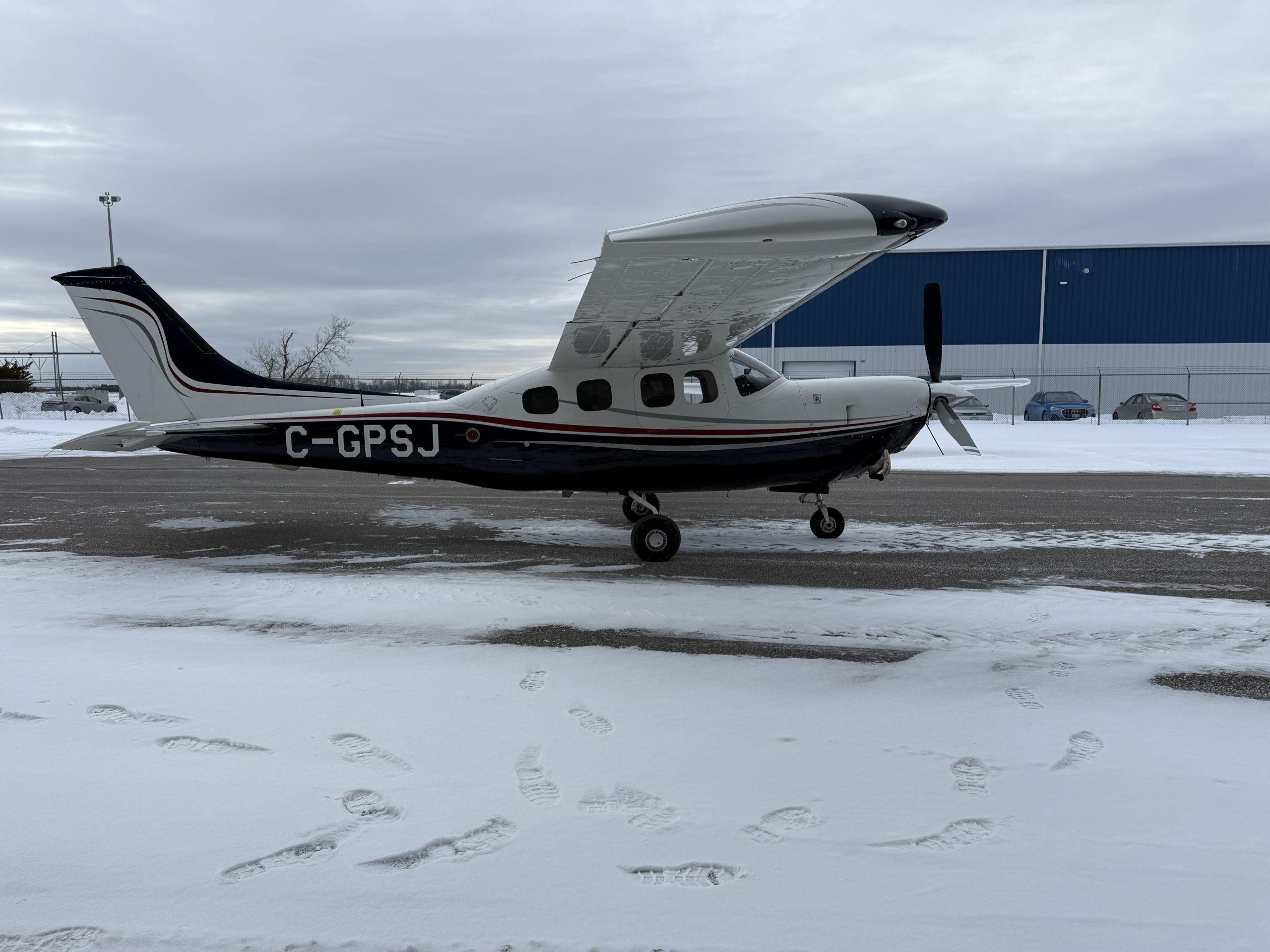 Small single engine propeller airplane parked on a snow covered airport tarmac with a blue hangar in the background