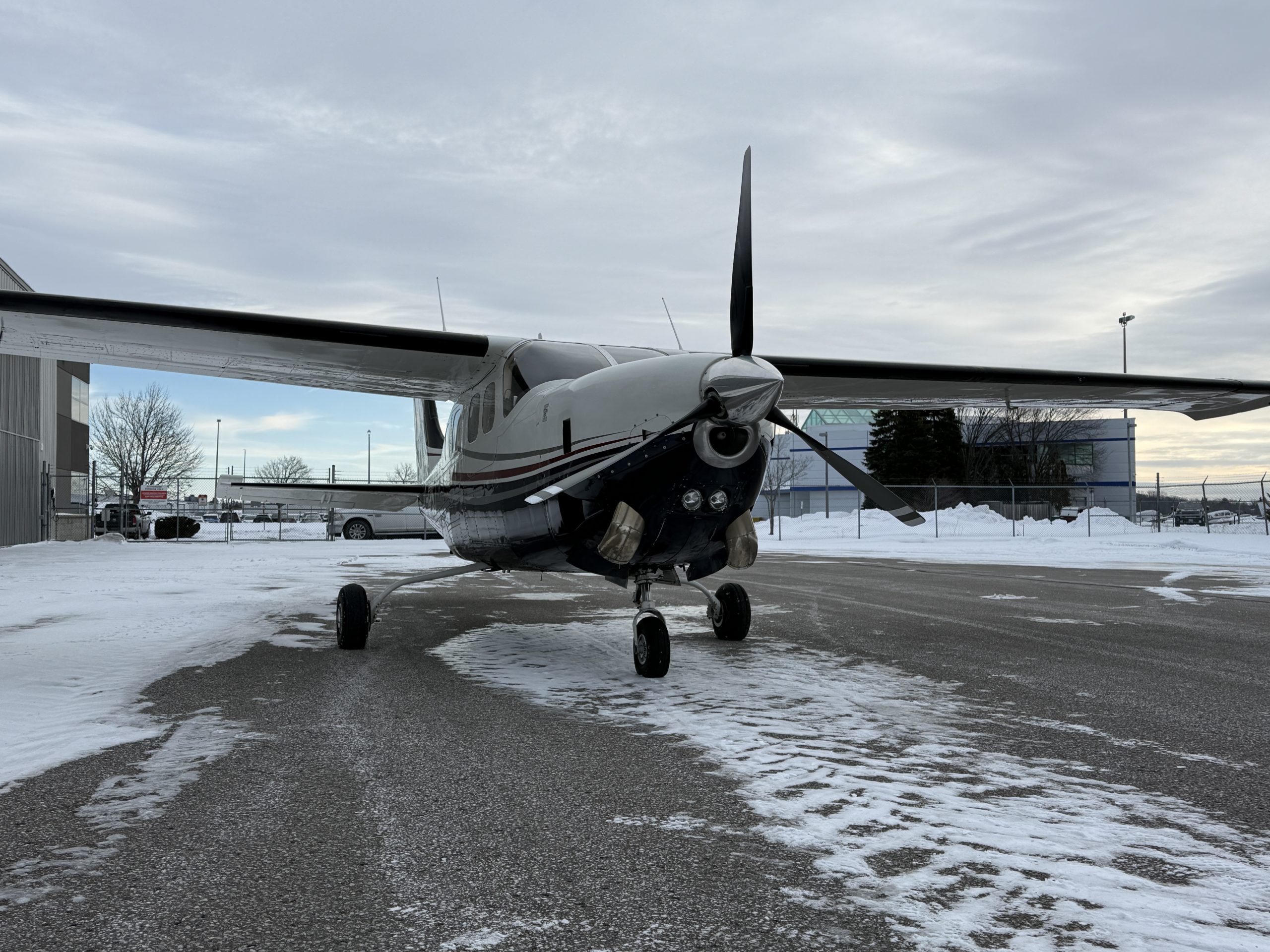 Twin engine propeller aircraft parked on a snow covered tarmac near a hangar with a fence and industrial buildings in the background