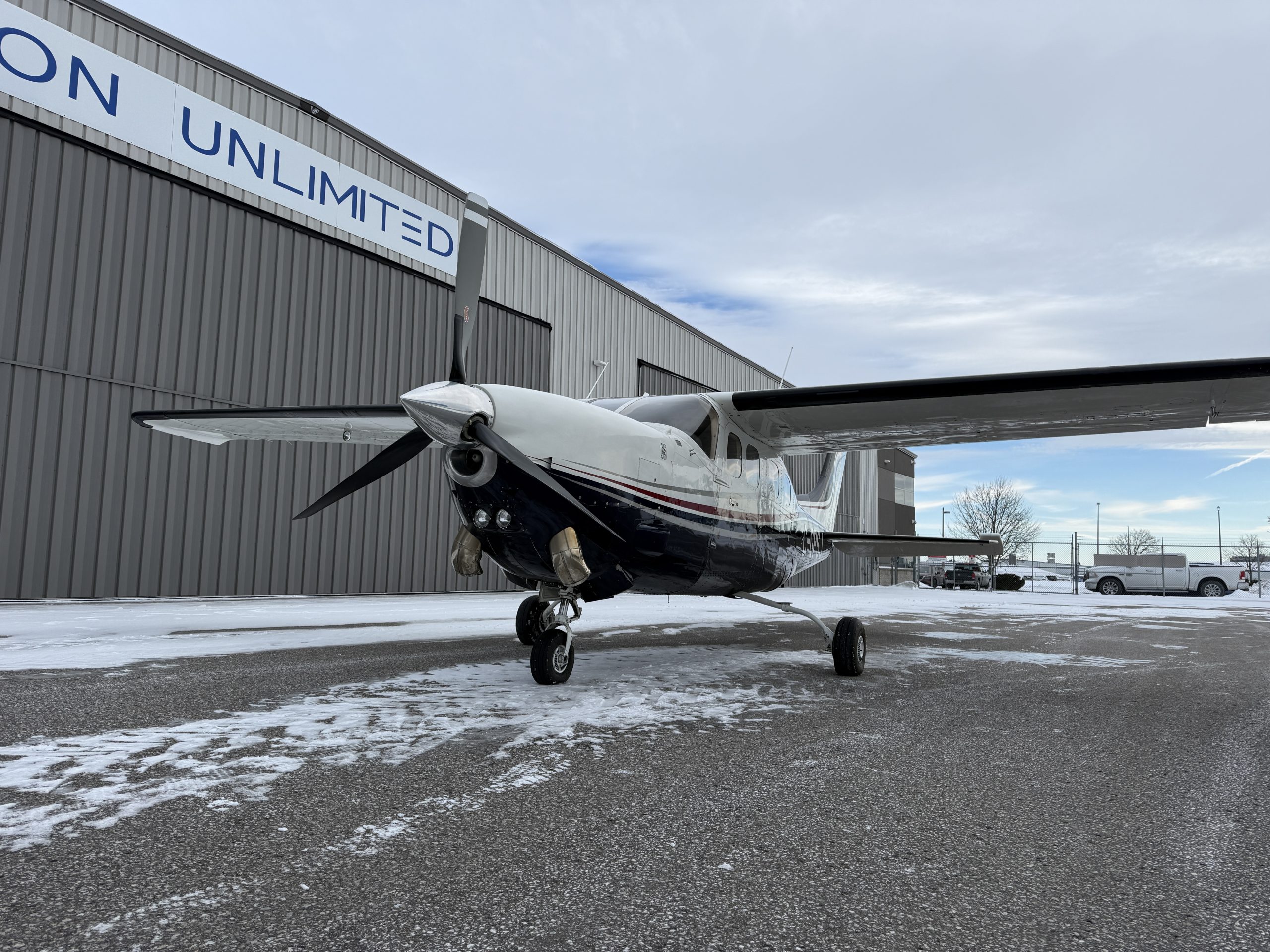 Propeller airplane parked on a snow covered tarmac beside a gray hangar with an UNLIMITED sign visible on the building