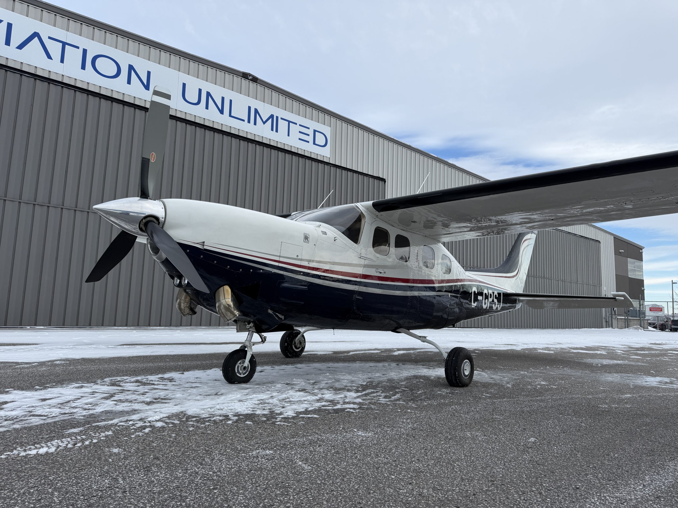 Small single engine propeller airplane parked on a snow dusted tarmac in front of a gray hangar with a large UNLIMITED sign overhead