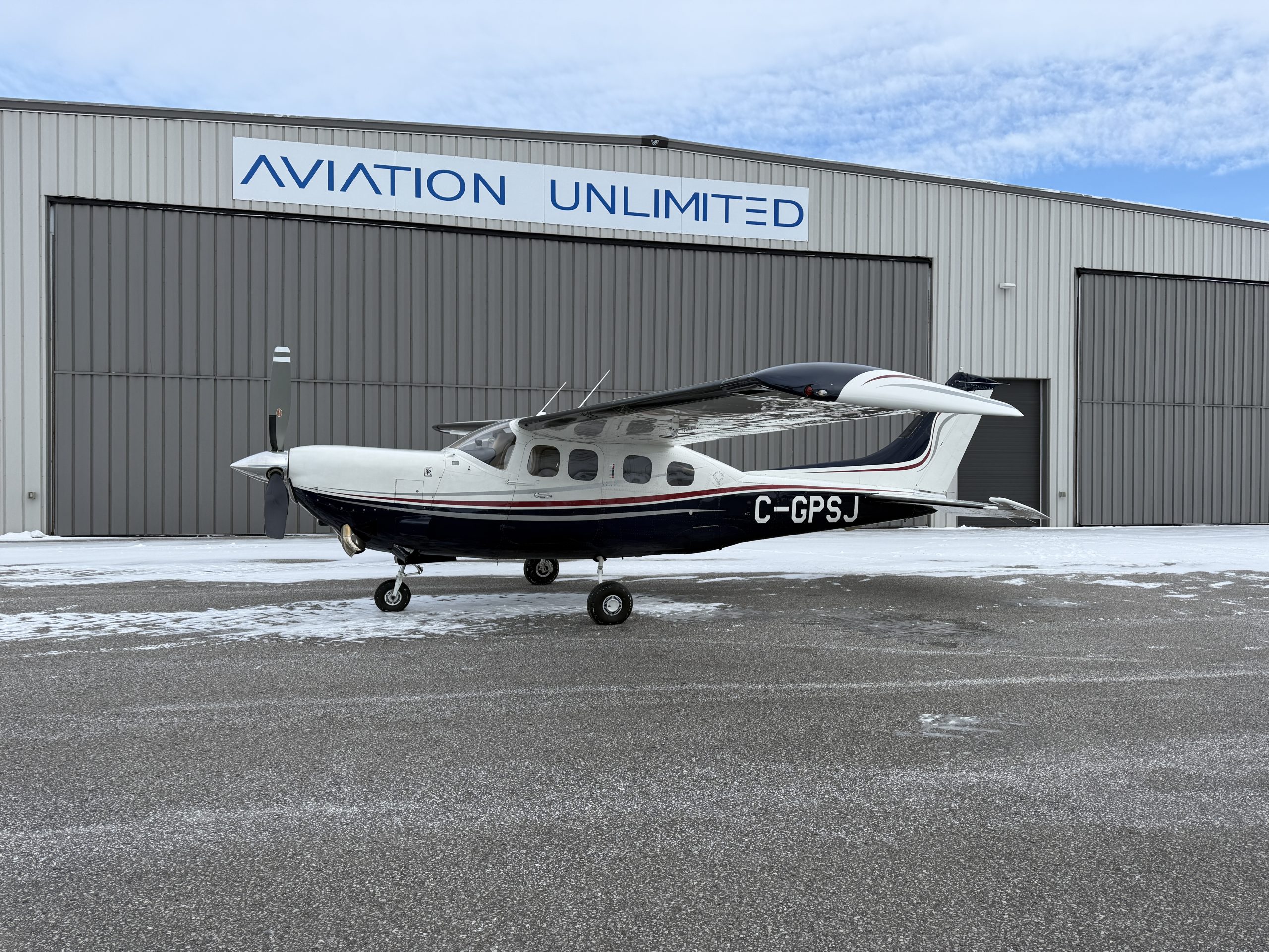 Small propeller airplane parked on a snowy tarmac in front of a gray hangar with a blue AVIATION UNLIMITED sign showing registration C GPSJ on the fuselage