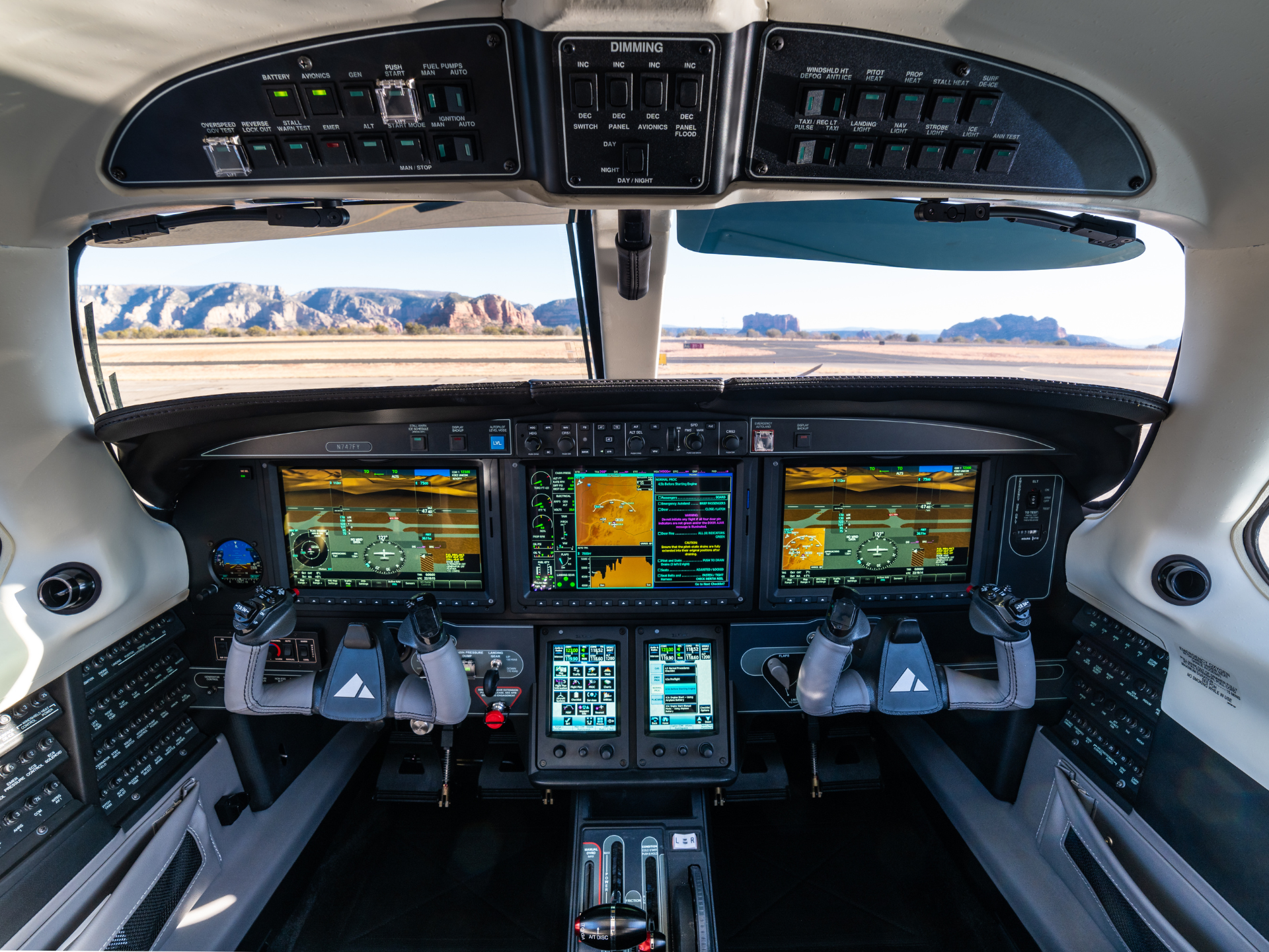 Modern aircraft cockpit with three large screens, flight controls, and overhead panels; desert landscape visible through windshield outside the windows.