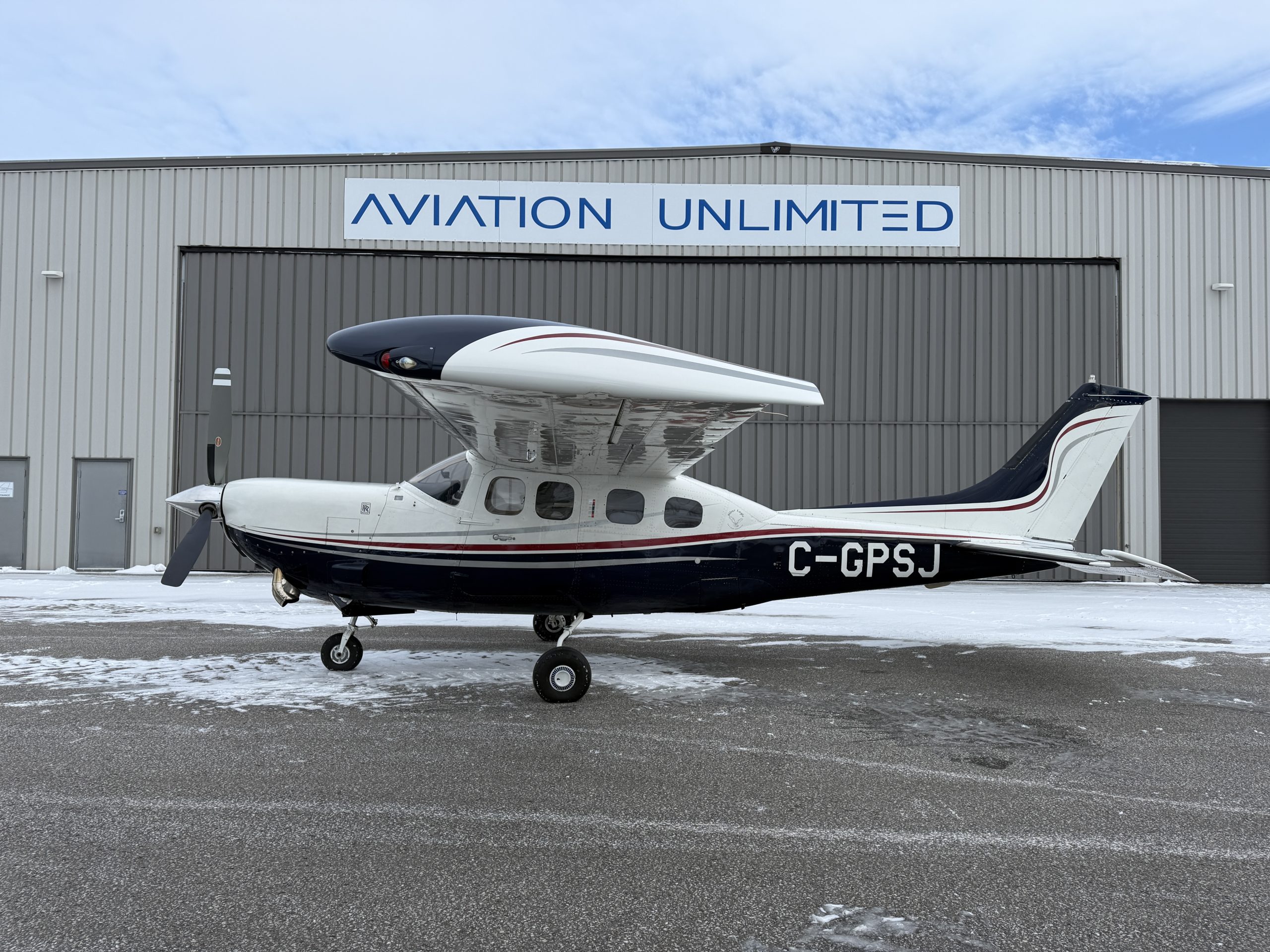 Small single engine airplane with white and dark blue accents parked on snow outside a hangar labeled Aviation Unlimited