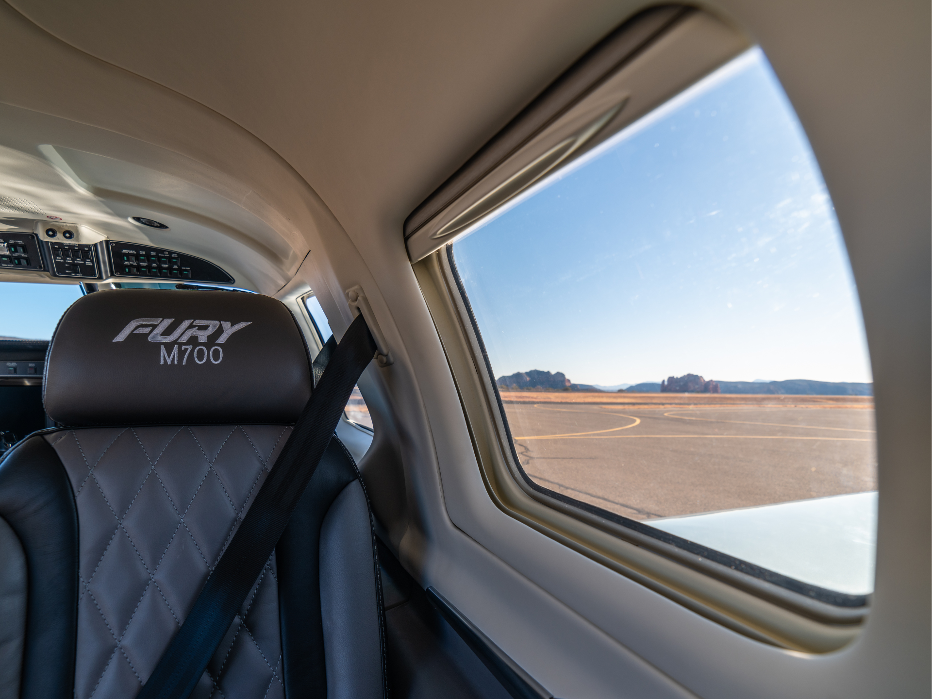 Cockpit of a small aircraft with a quilted black seat labeled 'FURY M700' and a window showing a runway and distant desert mountains.