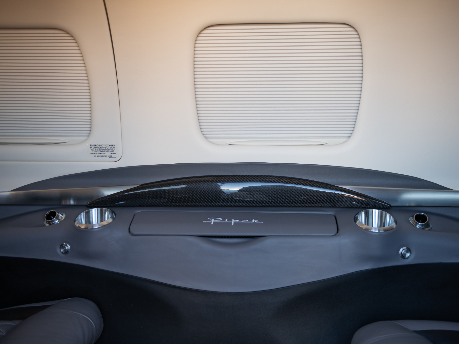 Luxury car interior viewed from the front, showing beige headliner, curved panels, and a carbon-fiber console with round cup holders