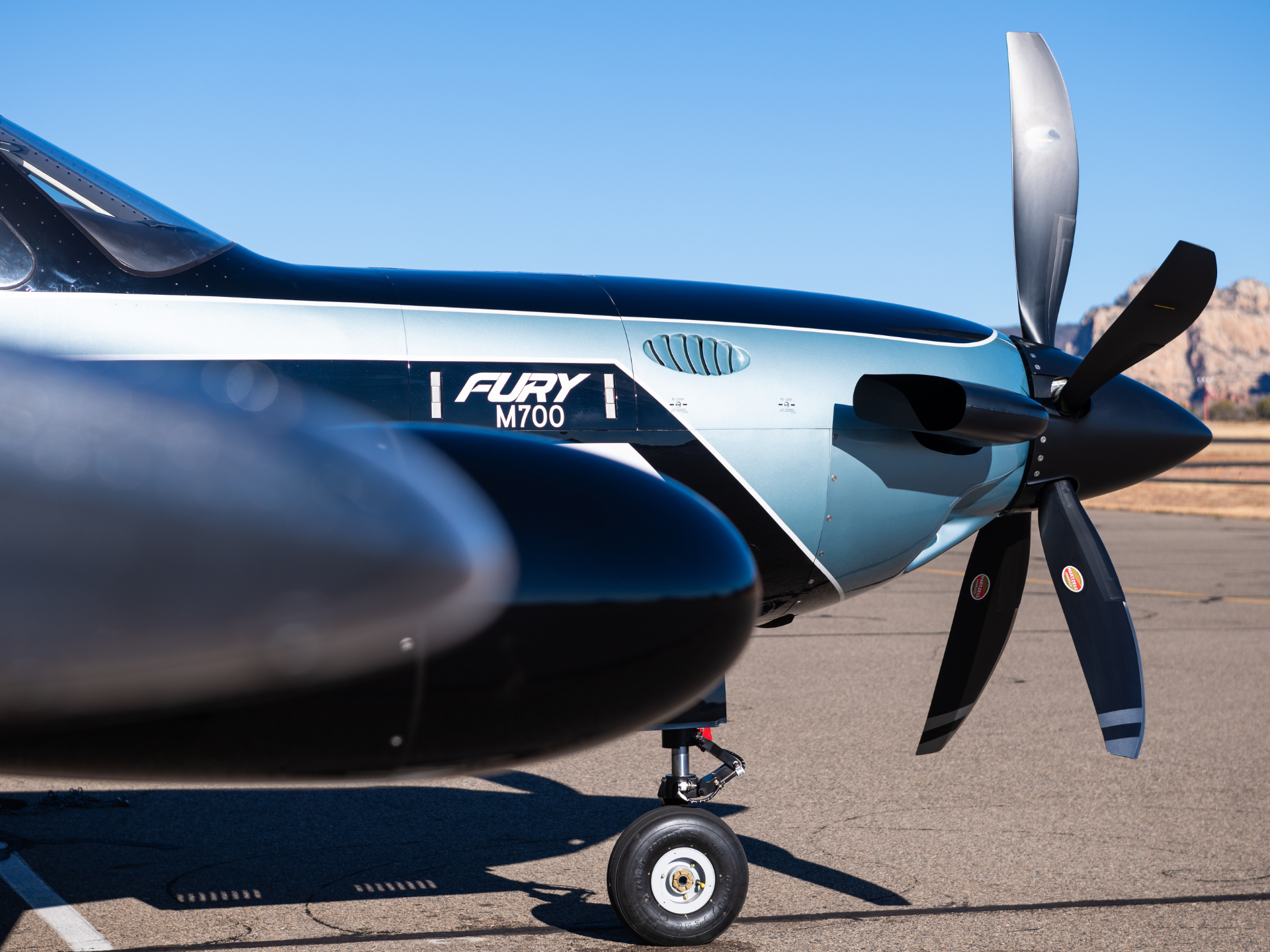 Close-up of a small propeller airplane with a three-blade propeller, Fury M700 branding, and nose wheel on a tarmac under a clear blue sky.