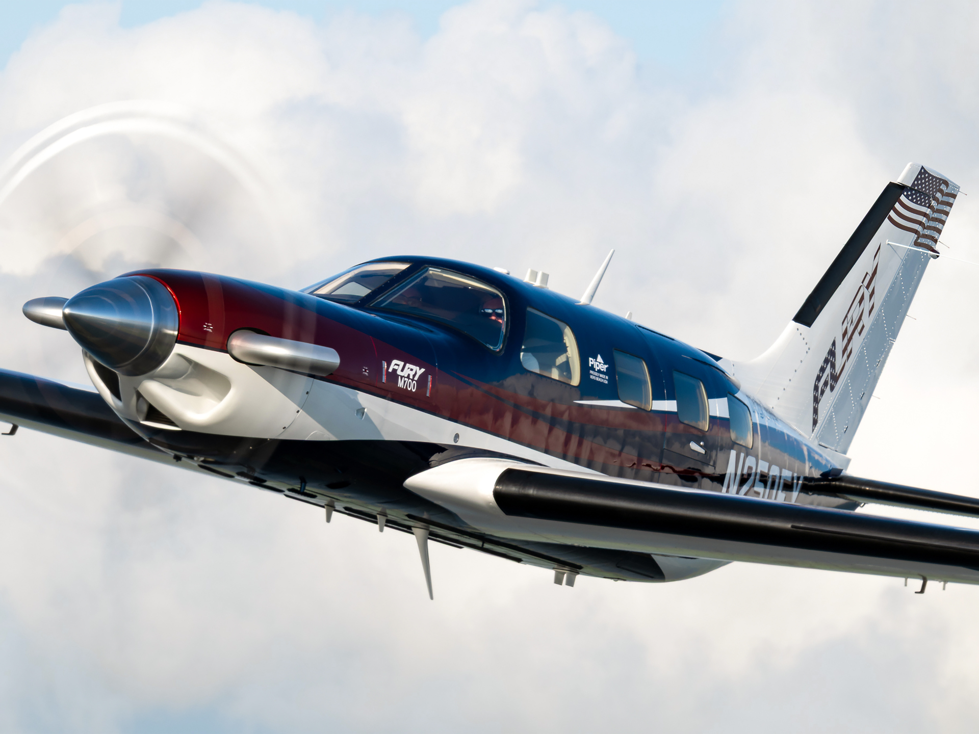 Blue, red, and white Piper M700 Fury aircraft in flight, propeller spinning, against a cloudy sky with an American flag on the tail.