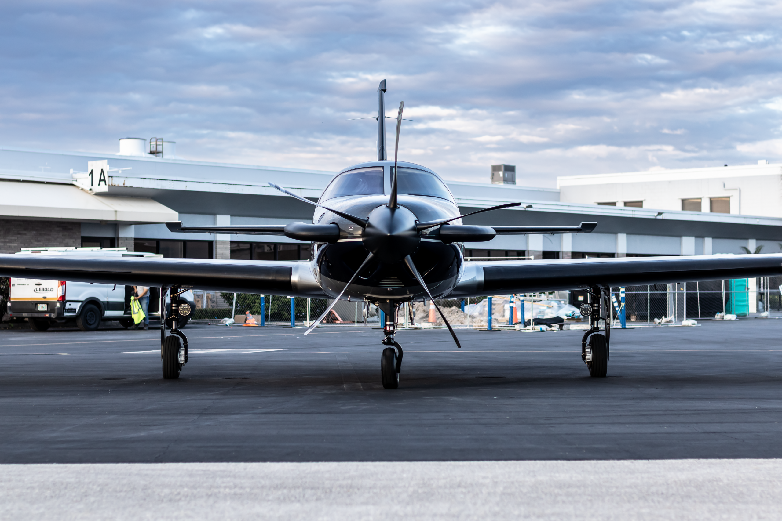 Front view of a small propeller airplane parked on a tarmac with a hangar in the background beneath a cloudy sky.